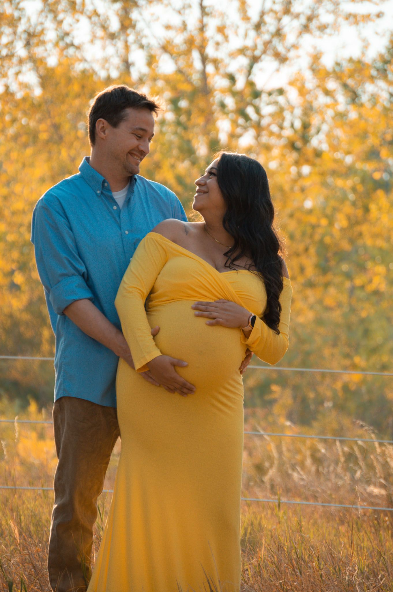 Smiling couple embracing pregnant belly in yellow dress among autumn foliage.