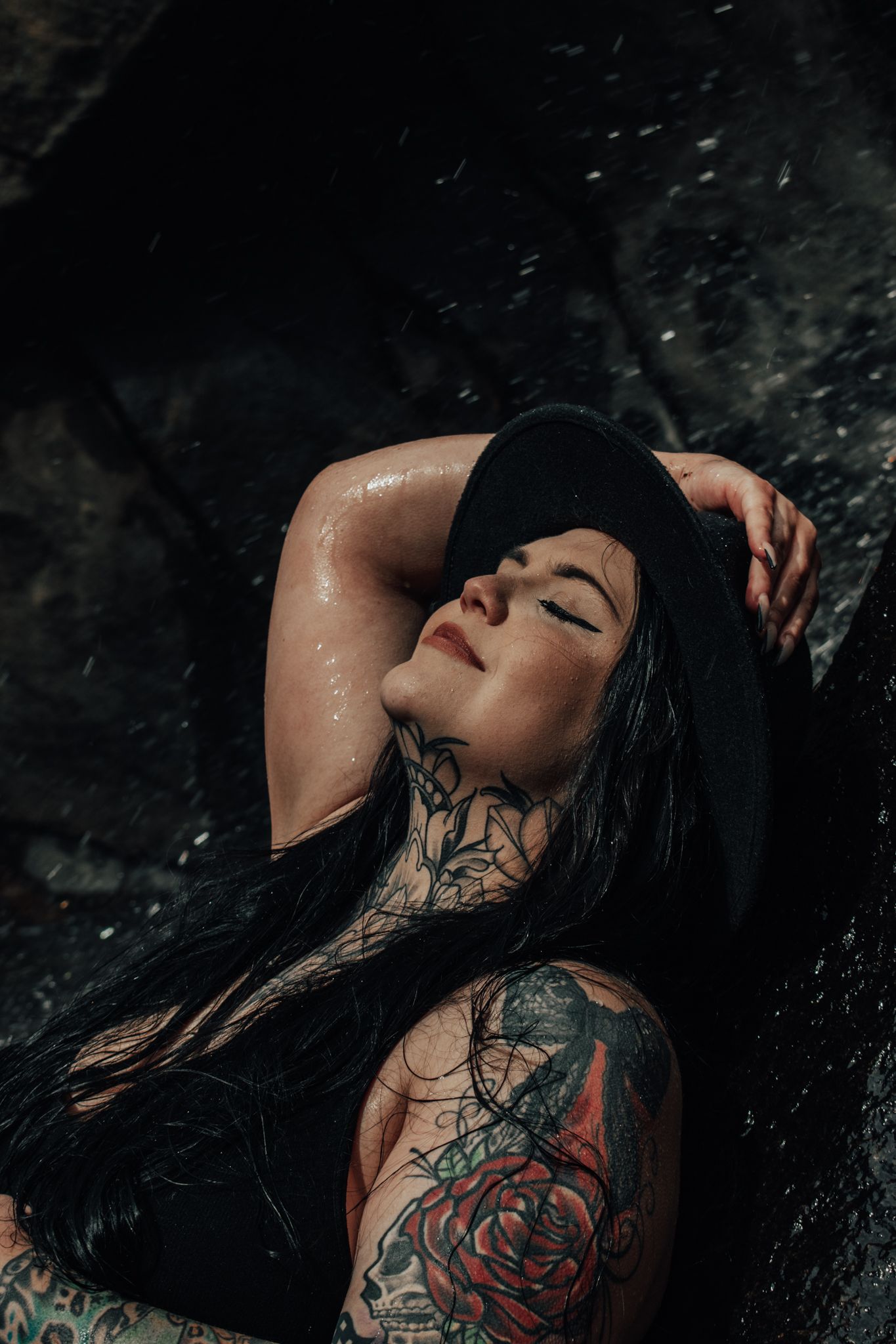 Tattooed woman with long dark hair lies on wet rocks with eyes closed in dramatic portrait.