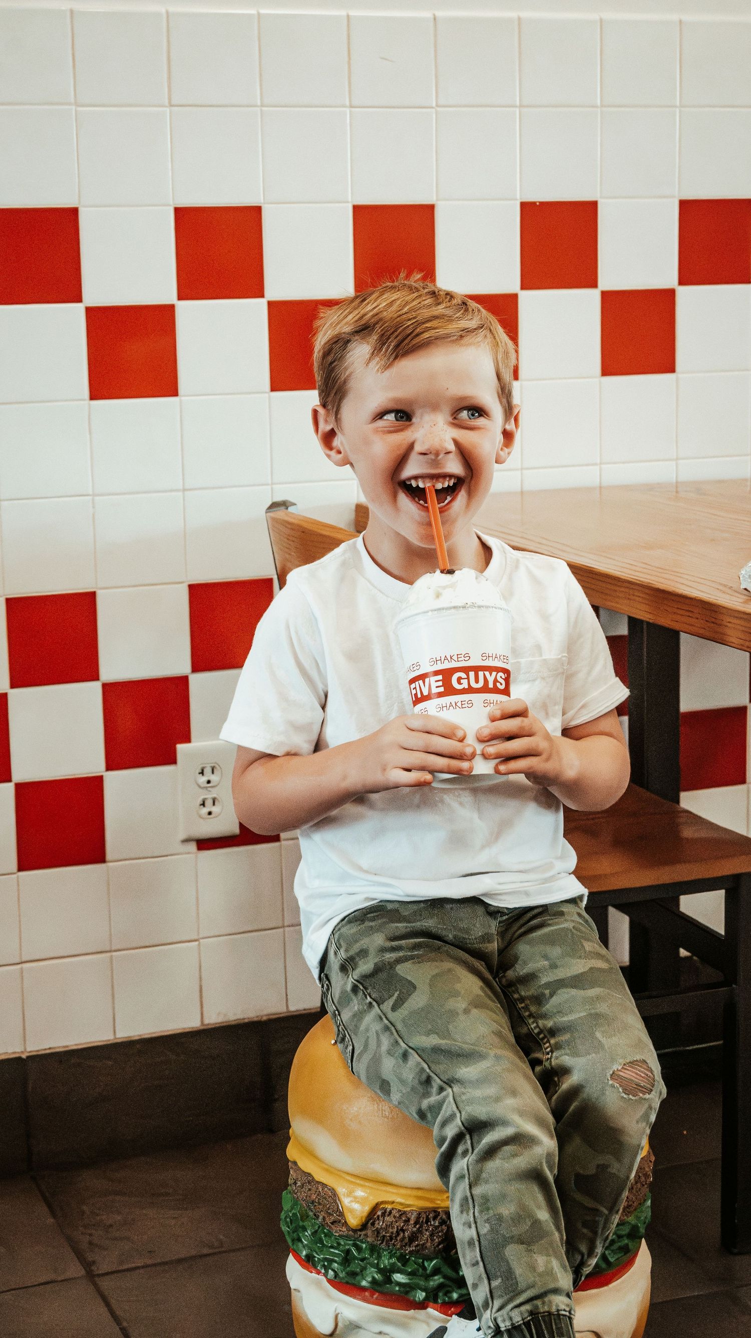 Cheerful young boy sitting on a burger stool holding a Five Guys drink cup in a restaurant.