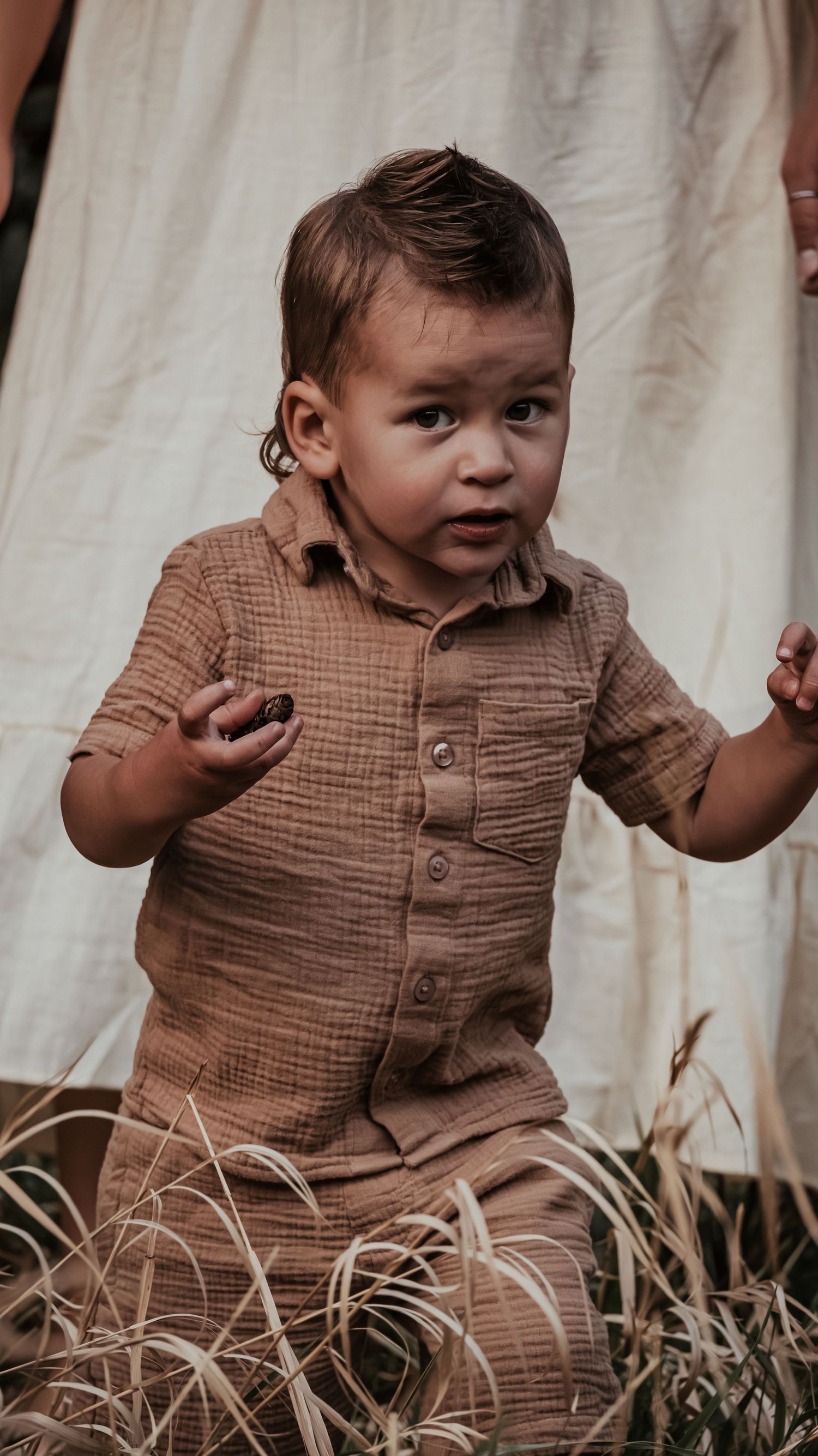 An adorable toddler boy in a brown linen outfit holds a small object while standing in a field.