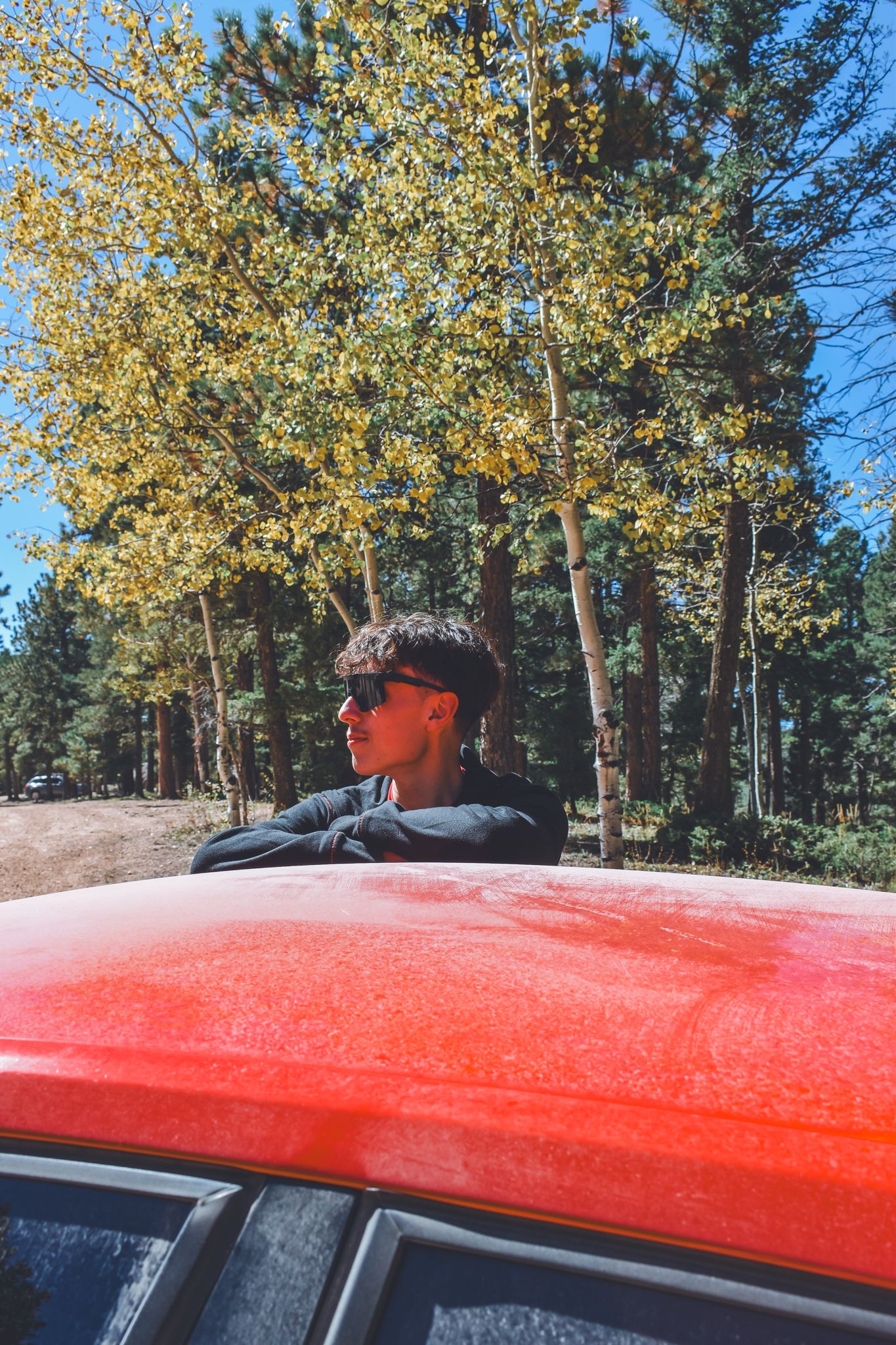 Young man in sunglasses leaning on a red car roof surrounded by autumn forest trees for senior photos.