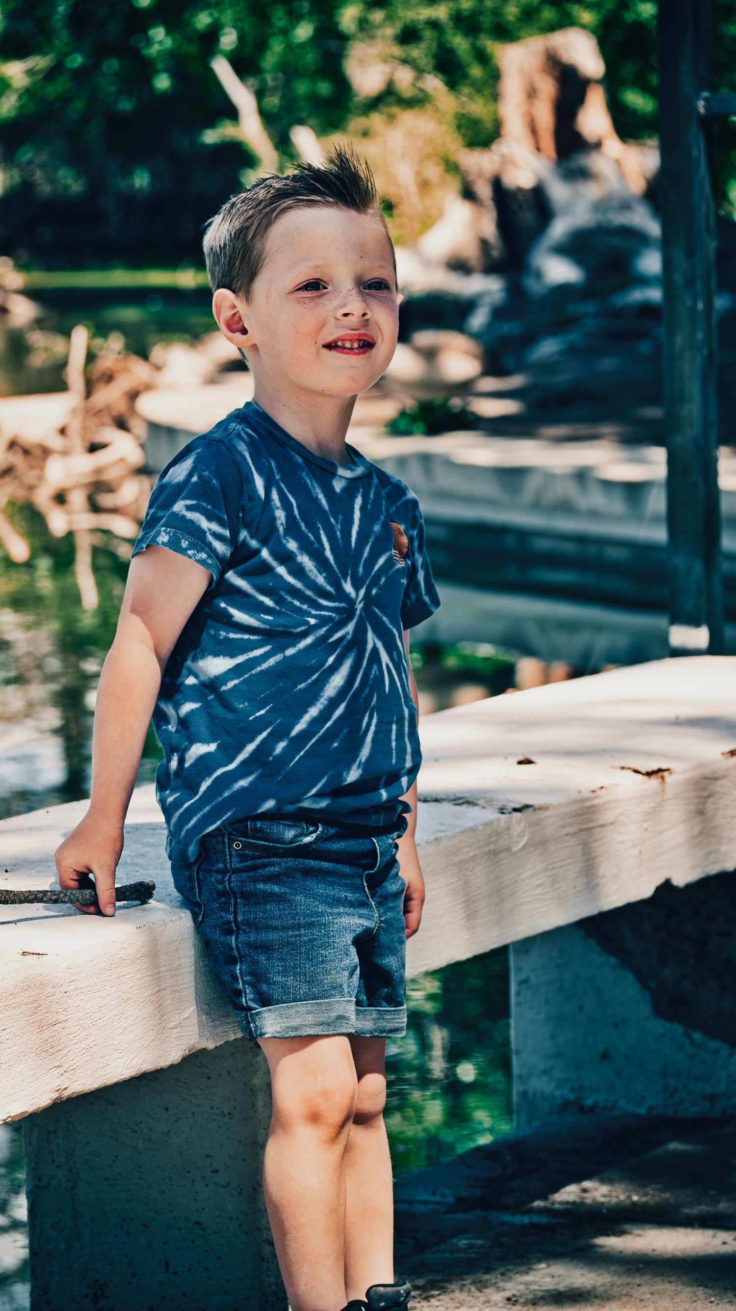 Young boy in blue tie-dye shirt and denim shorts leans on concrete railing near water. Family Photographer in Pflugerville, Tx