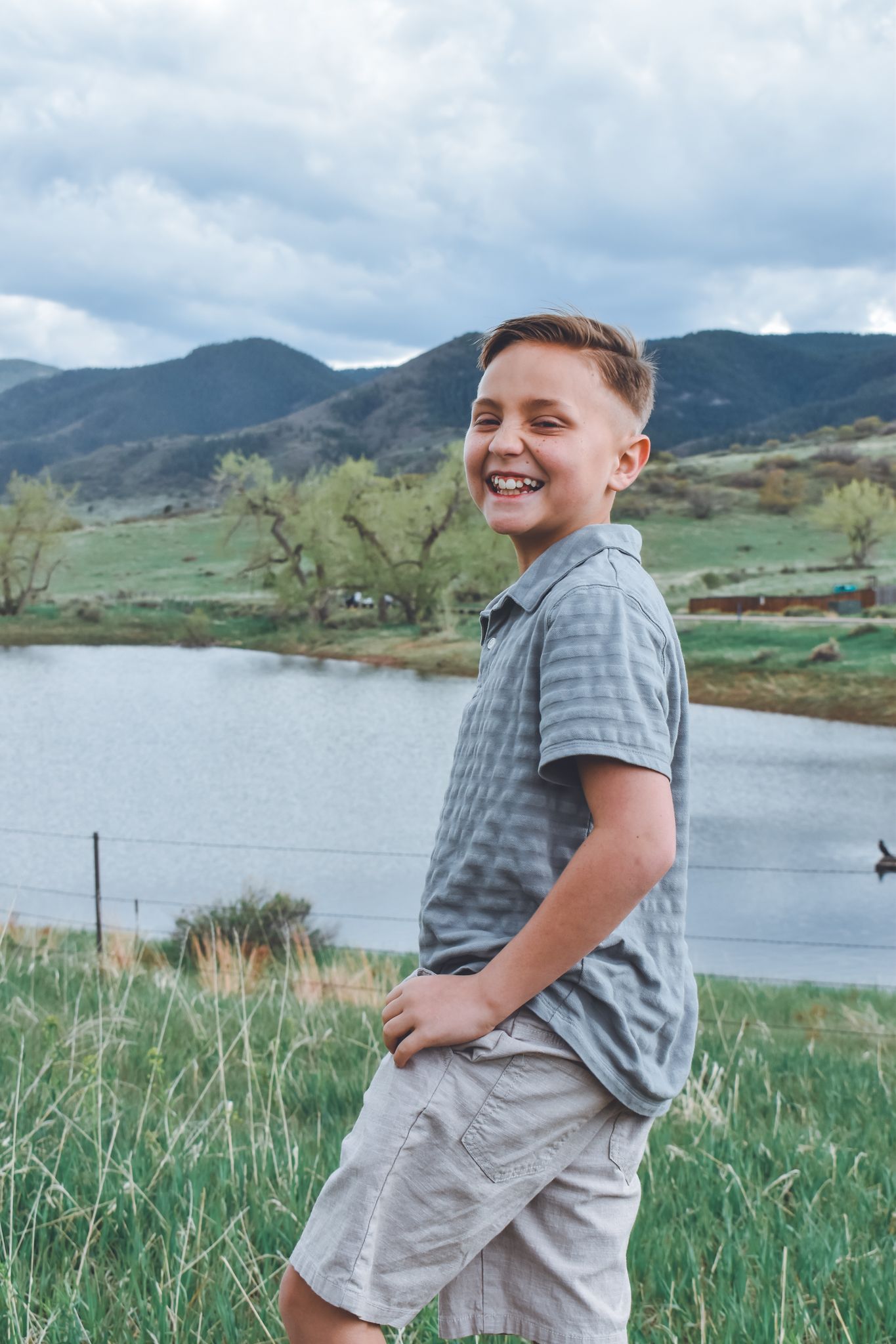 Smiling preteen boy in gray polo shirt and khaki shorts standing near mountain lake. Family Photographer in Pflugerville, Tx