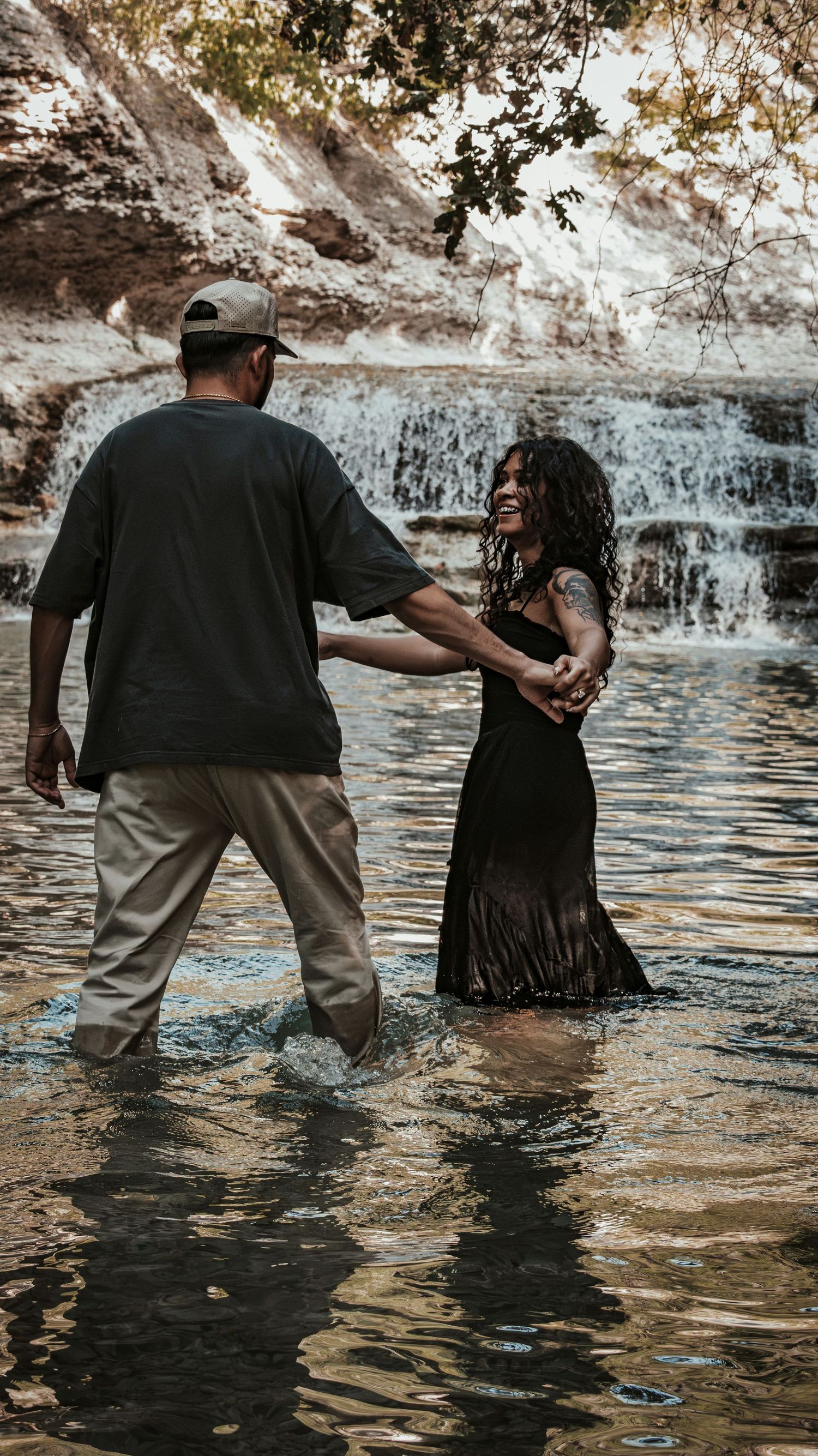 Couple holding hands wading through shallow water near a cascading waterfall in nature for a couples session.