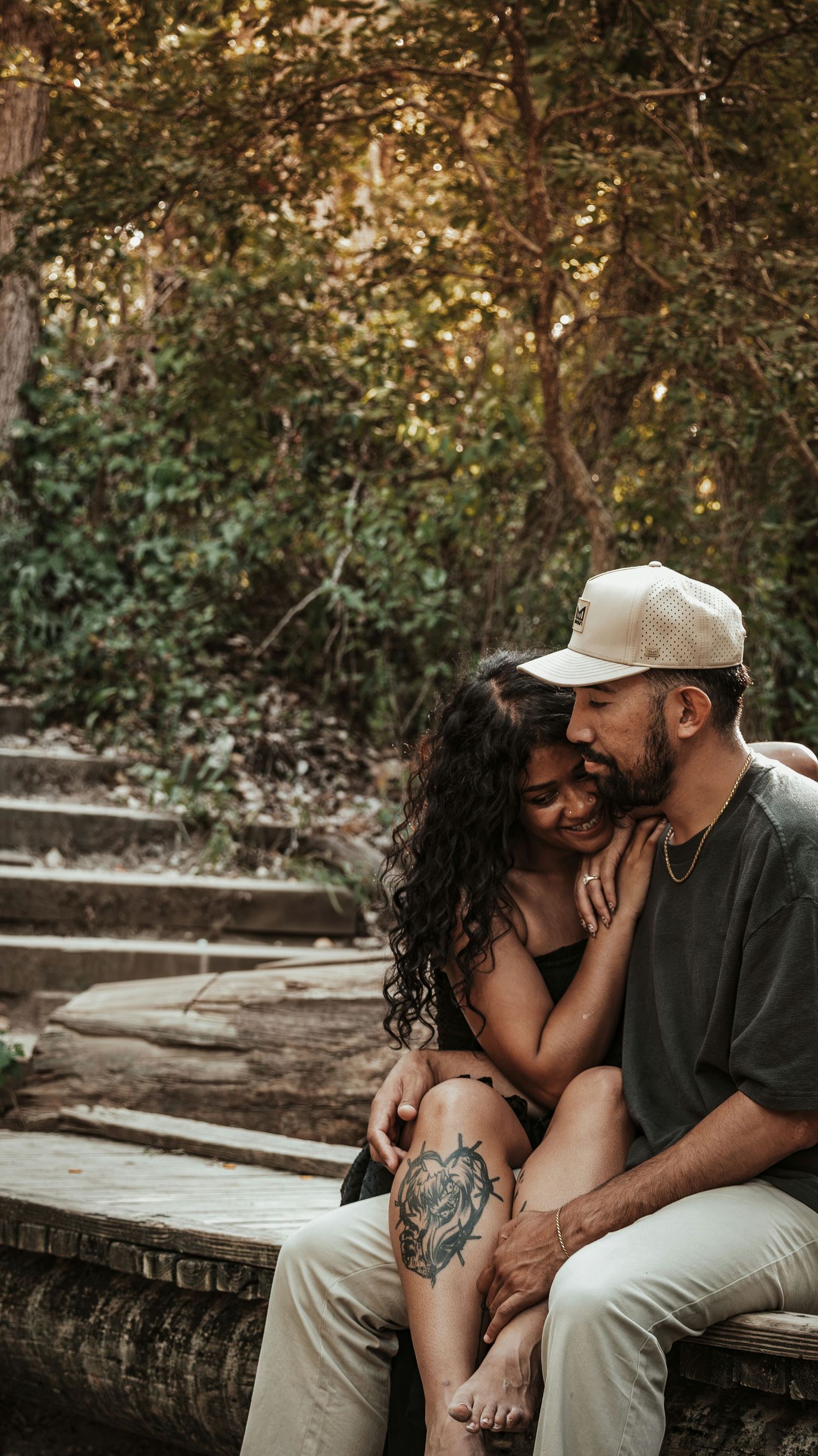 Happy couple cuddling on stone steps surrounded by trees, woman with tattoo leaning into mans embrace.