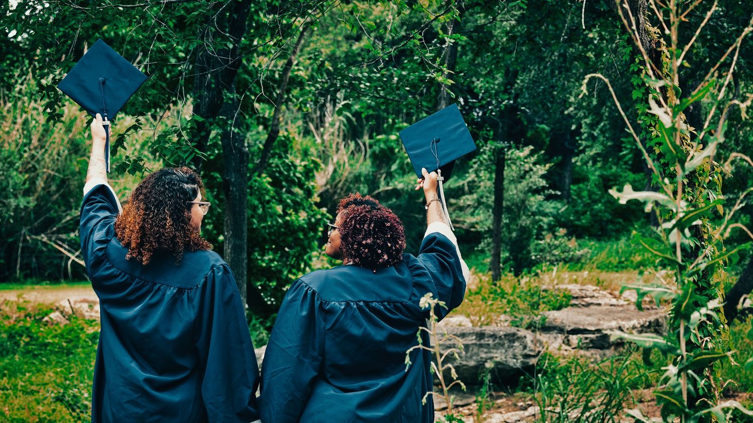 Two graduates in black robes raise mortarboard caps triumphantly in a lush green forest for senior photos.