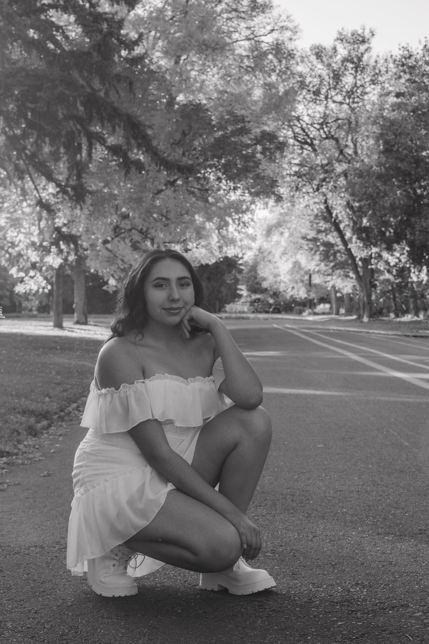Black and white portrait of young woman in off-shoulder dress crouching on park road for senior photos.