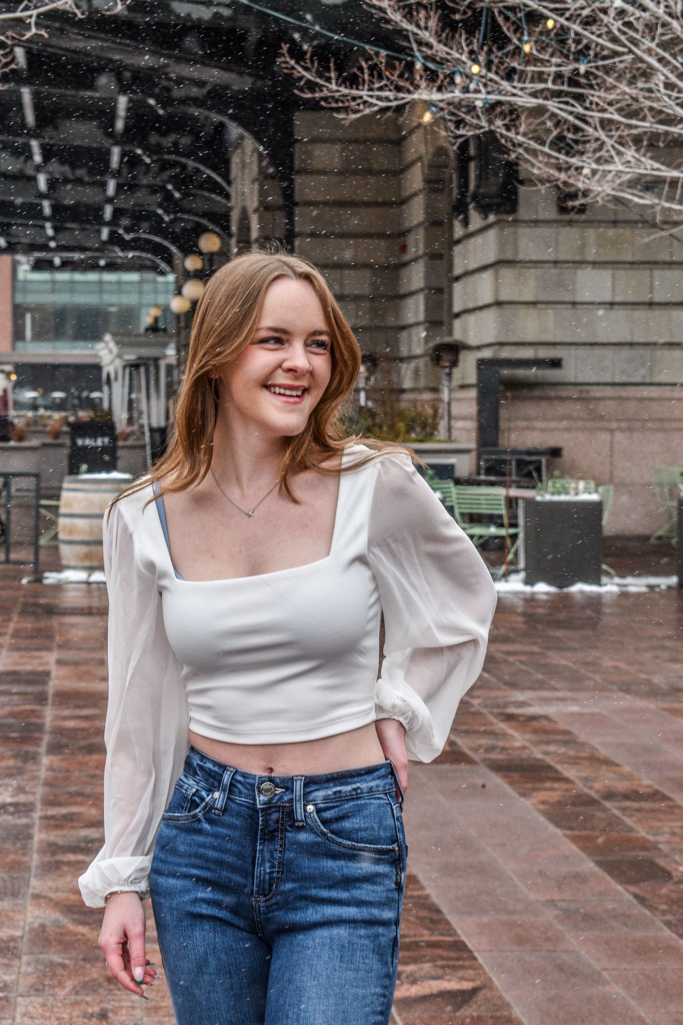 Smiling young woman in white square-neck crop top and jeans posing in snowy urban outdoor setting for senior photos.