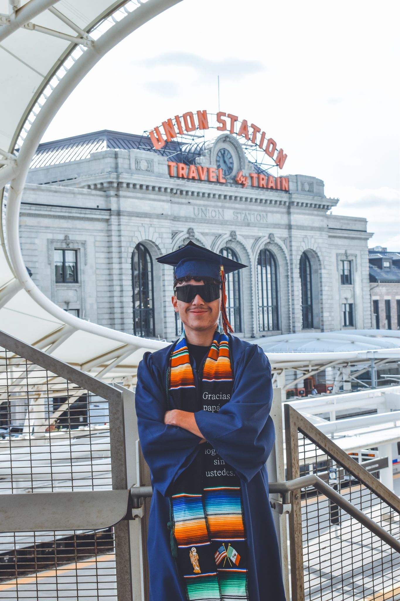 Graduate in blue cap and gown stands proudly in front of Denver Union Station landmark for senior photos.