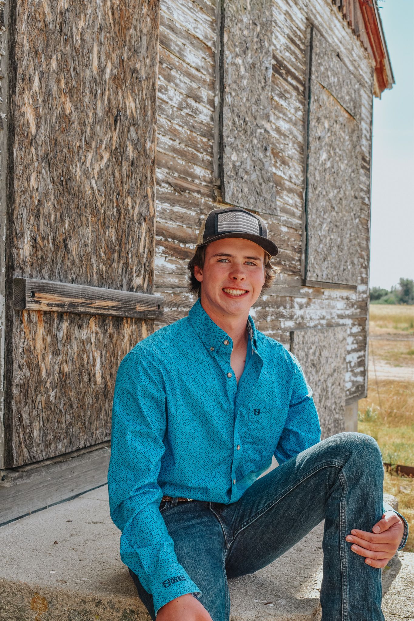 Smiling young man in teal western shirt and cap sitting on steps beside a rustic weathered barn for senior photos.