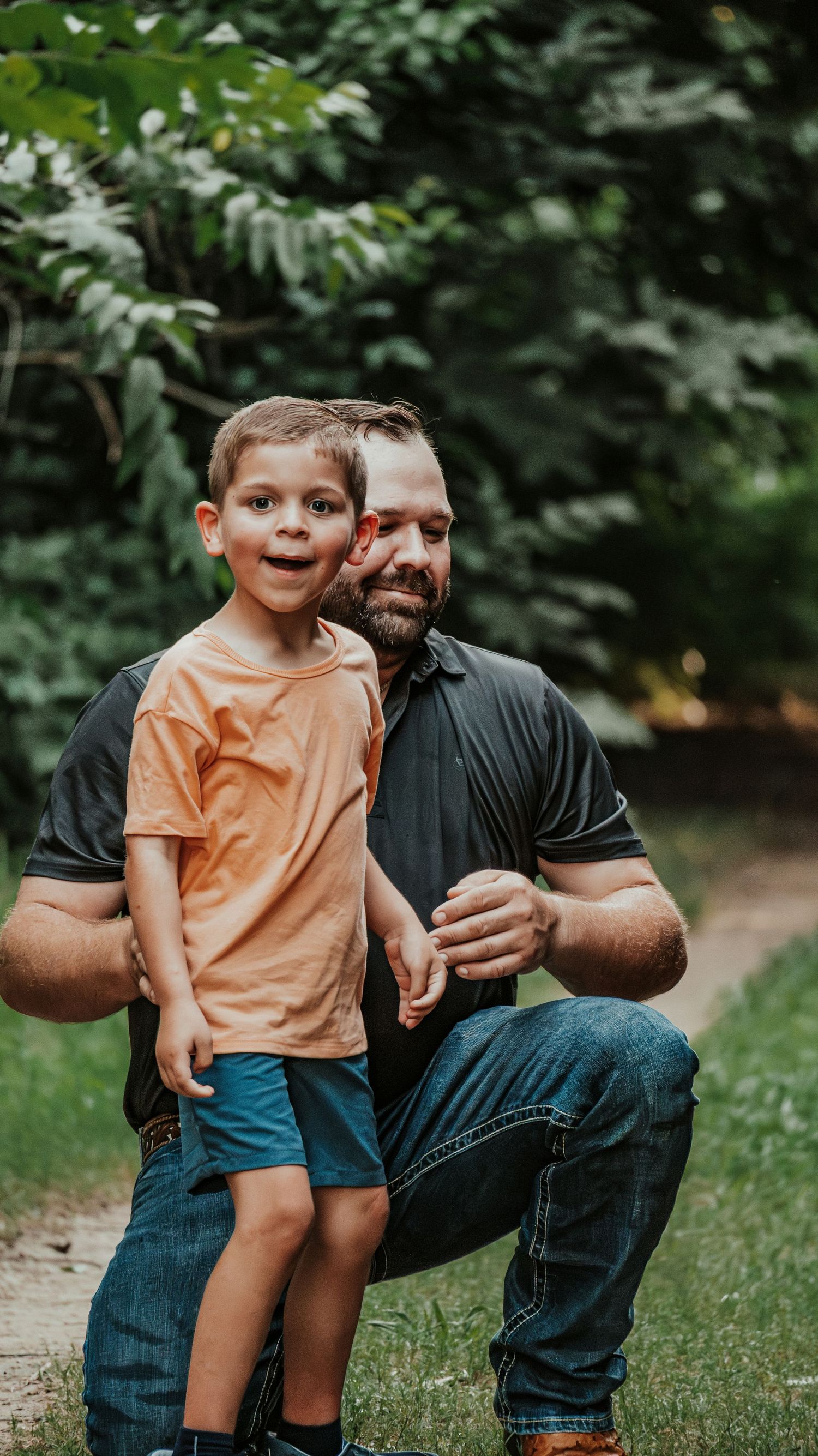 Bearded father crouching beside his young son on a lush green park path.