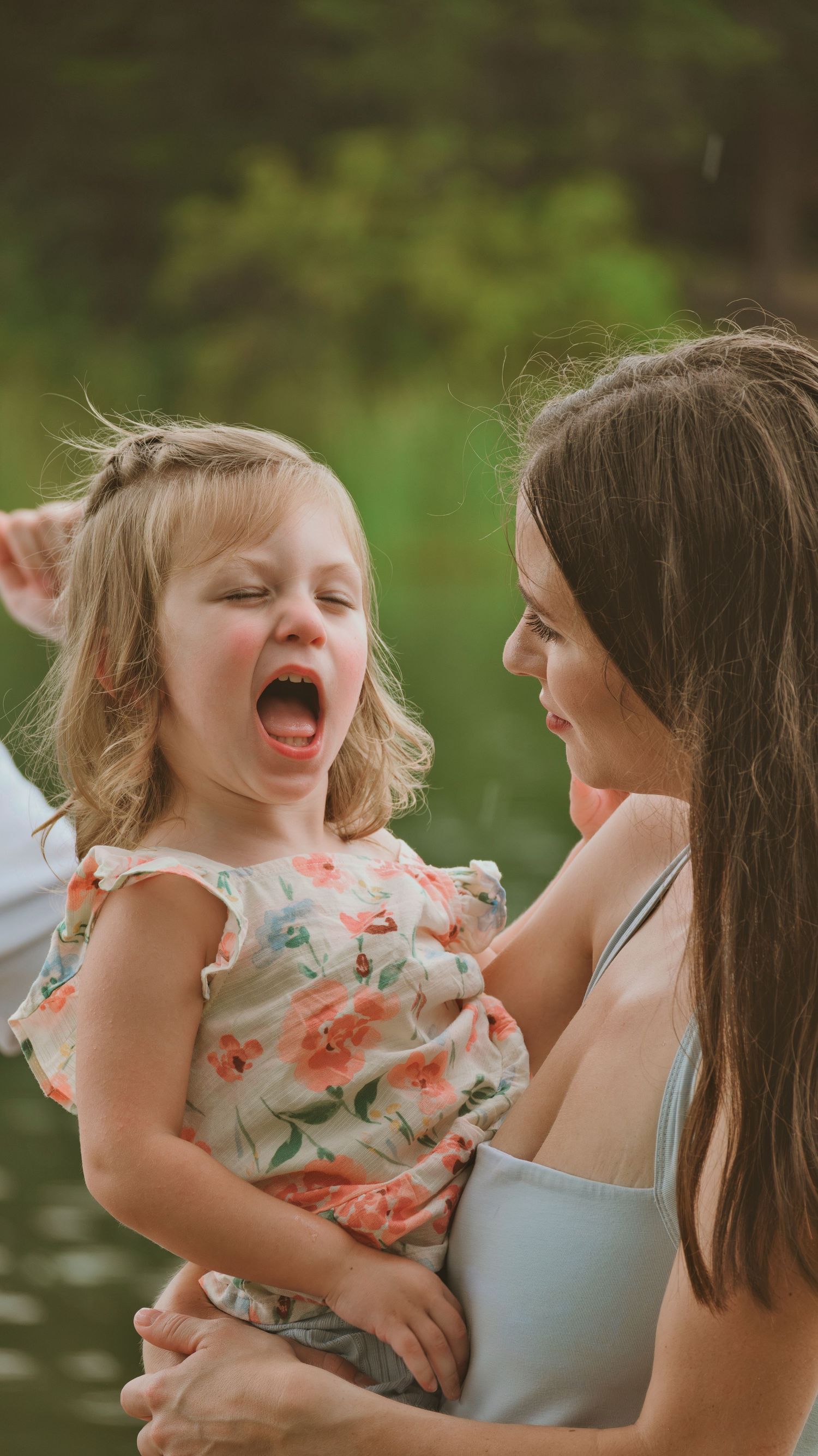Laughing toddler girl in floral dress being held by her smiling brunette mother outdoors.