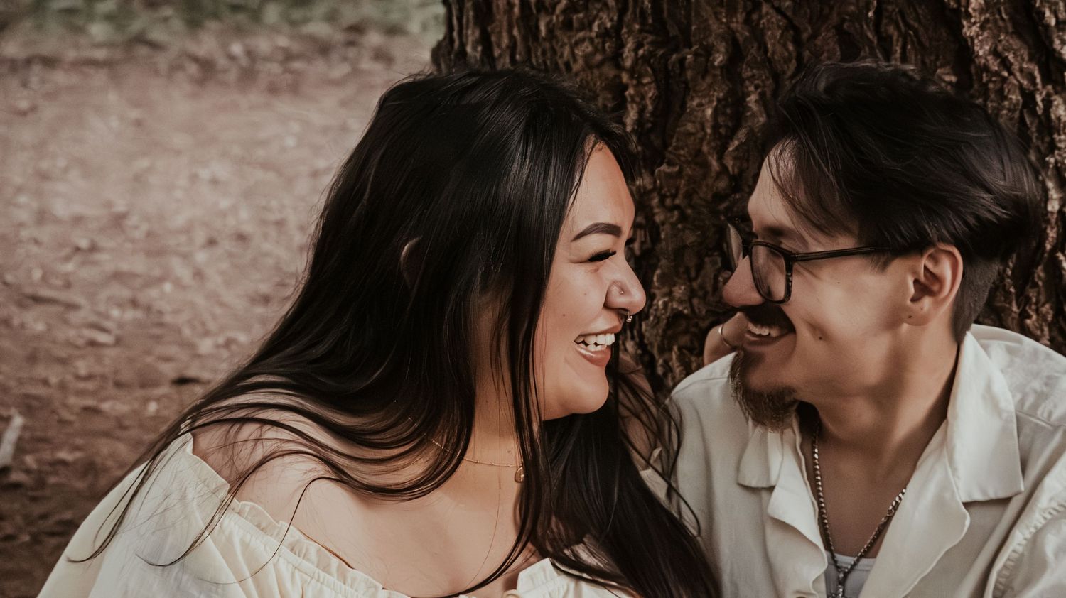 A couple laughs and smiles at each other while sitting near a tree outdoors.