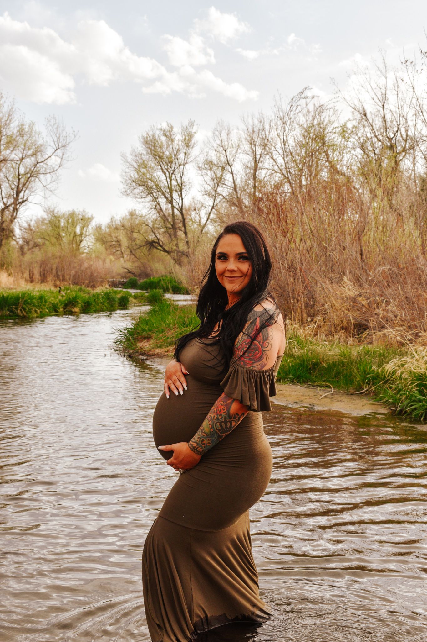 Smiling tattooed pregnant woman in olive maternity dress standing in shallow stream outdoors.