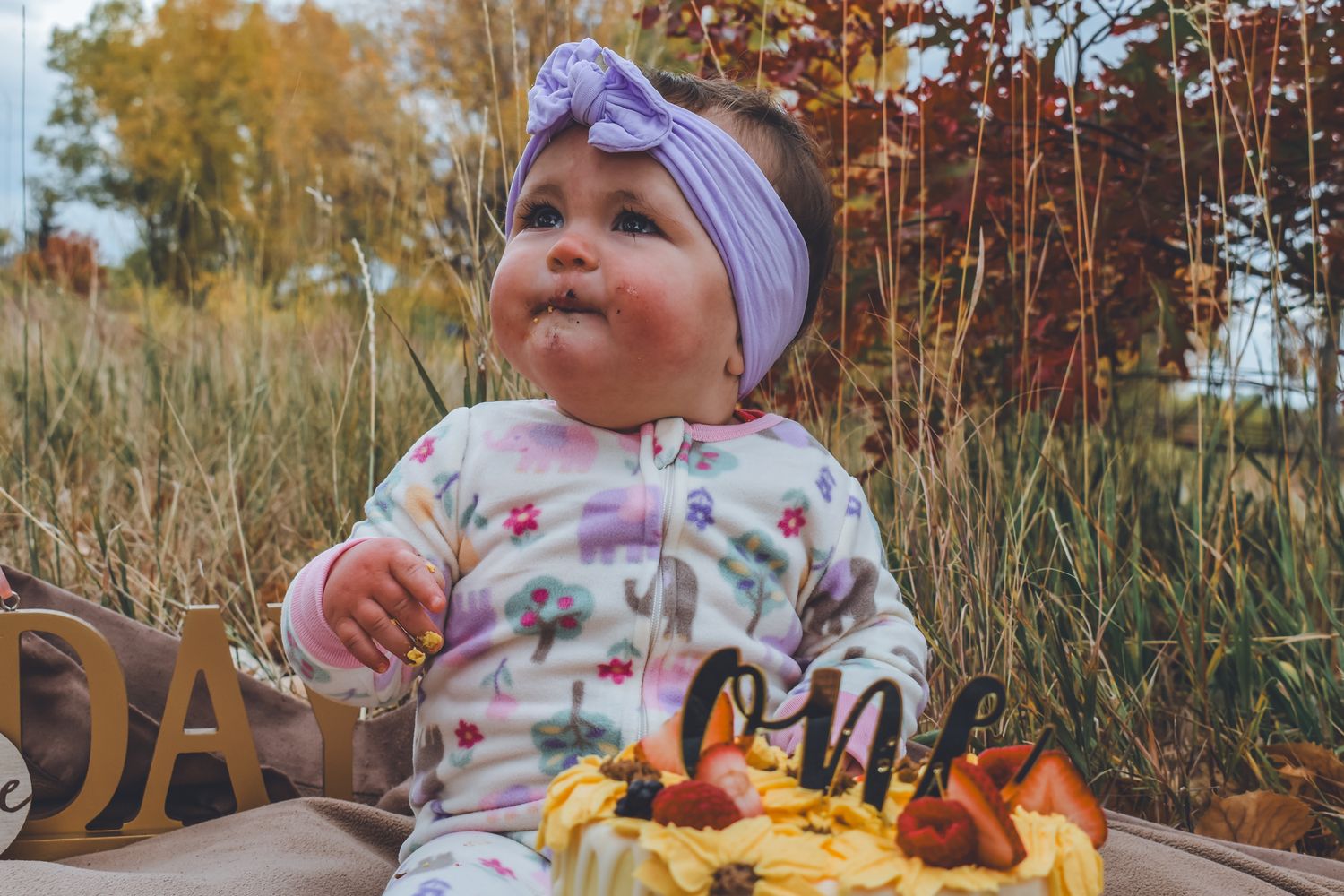 An adorable baby girl sits beside a smash cake during her first birthday celebration outdoors. Family Photographer in Pflugerville, Tx