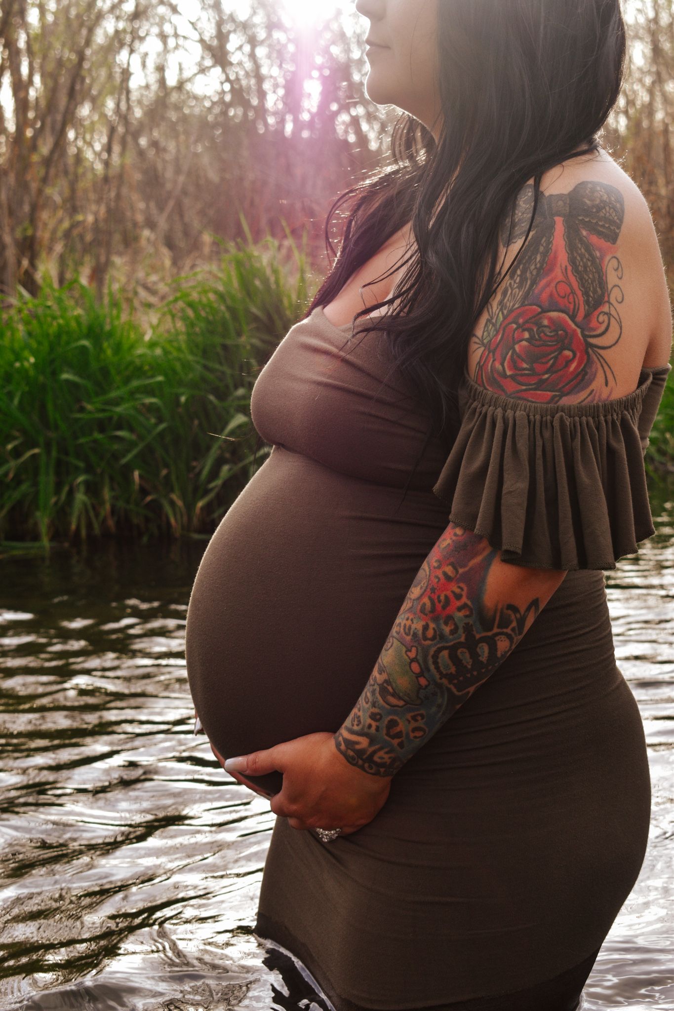 Pregnant woman with tattoos in brown dress standing in water surrounded by lush greenery. Maternity Photographer in Pflugerville, Tx