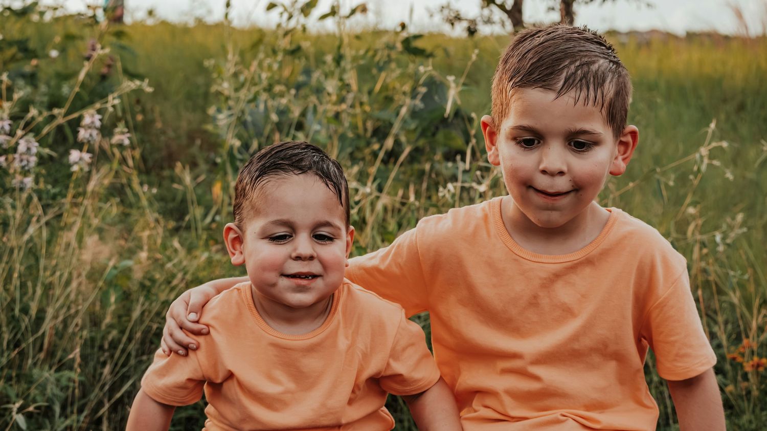 Two young brothers in matching orange shirts smile together while sitting in a sunny wildflower meadow.