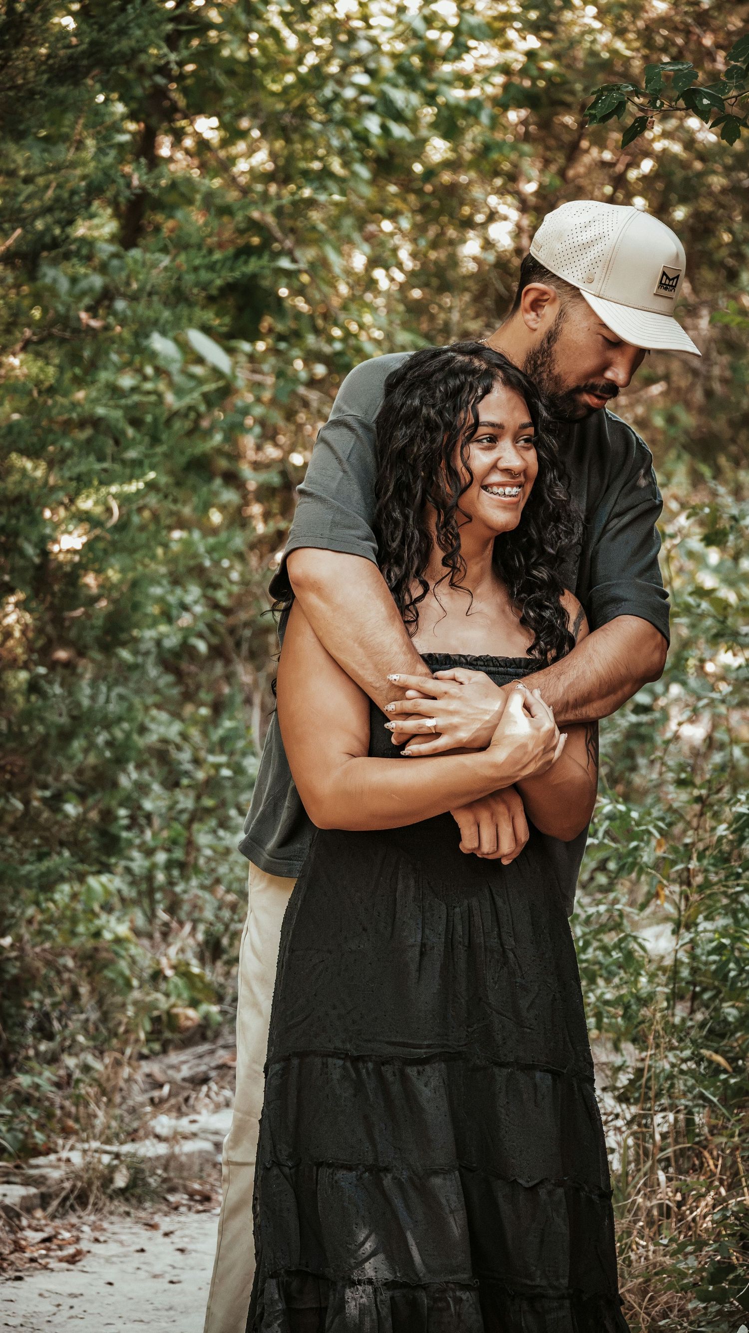 Man in white cap hugging woman in black maxi dress from behind on a wooded path, both smiling joyfully.