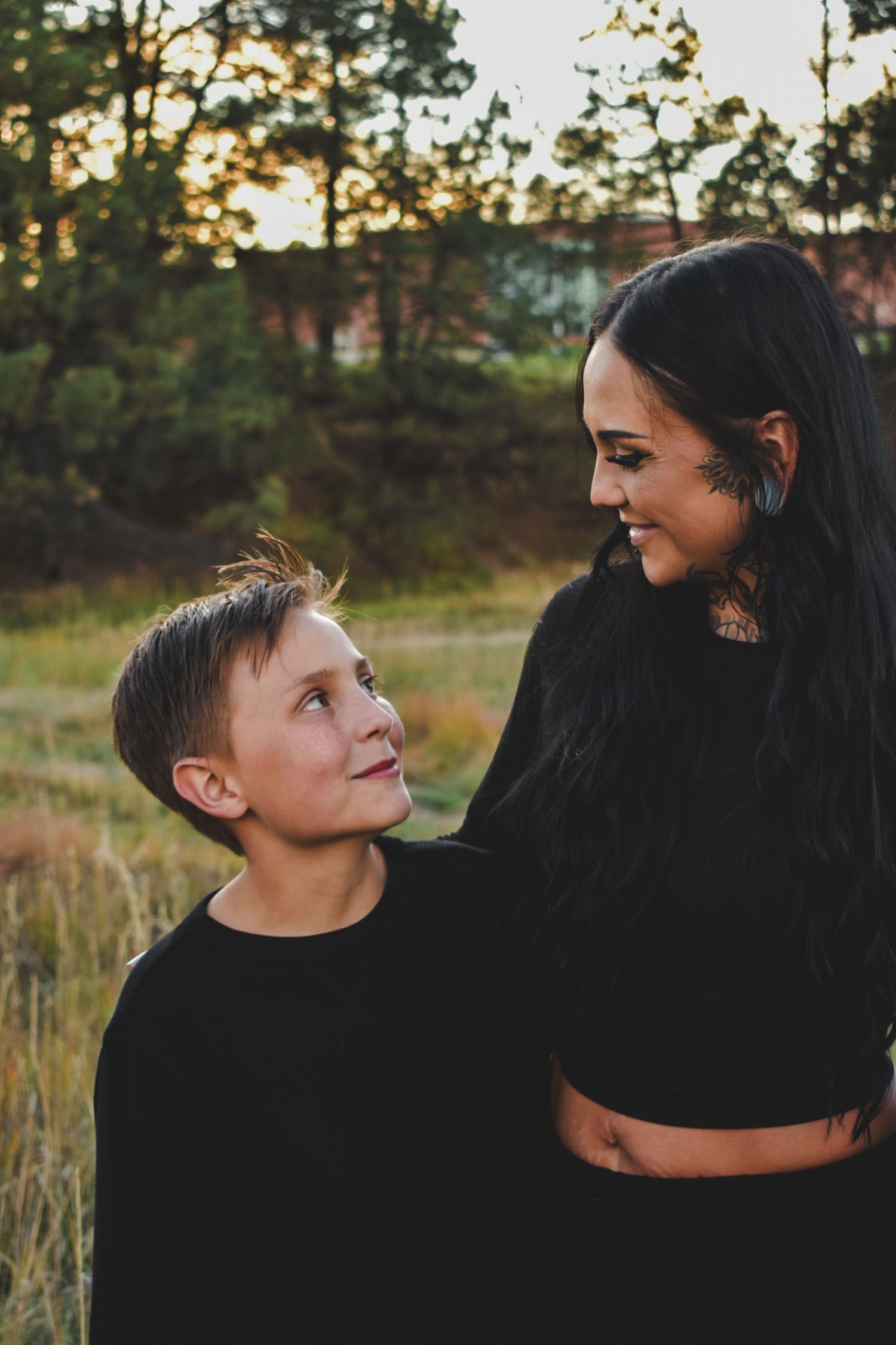 A young boy in black looks up adoringly at his tattooed mother during a golden hour outdoor photo session.