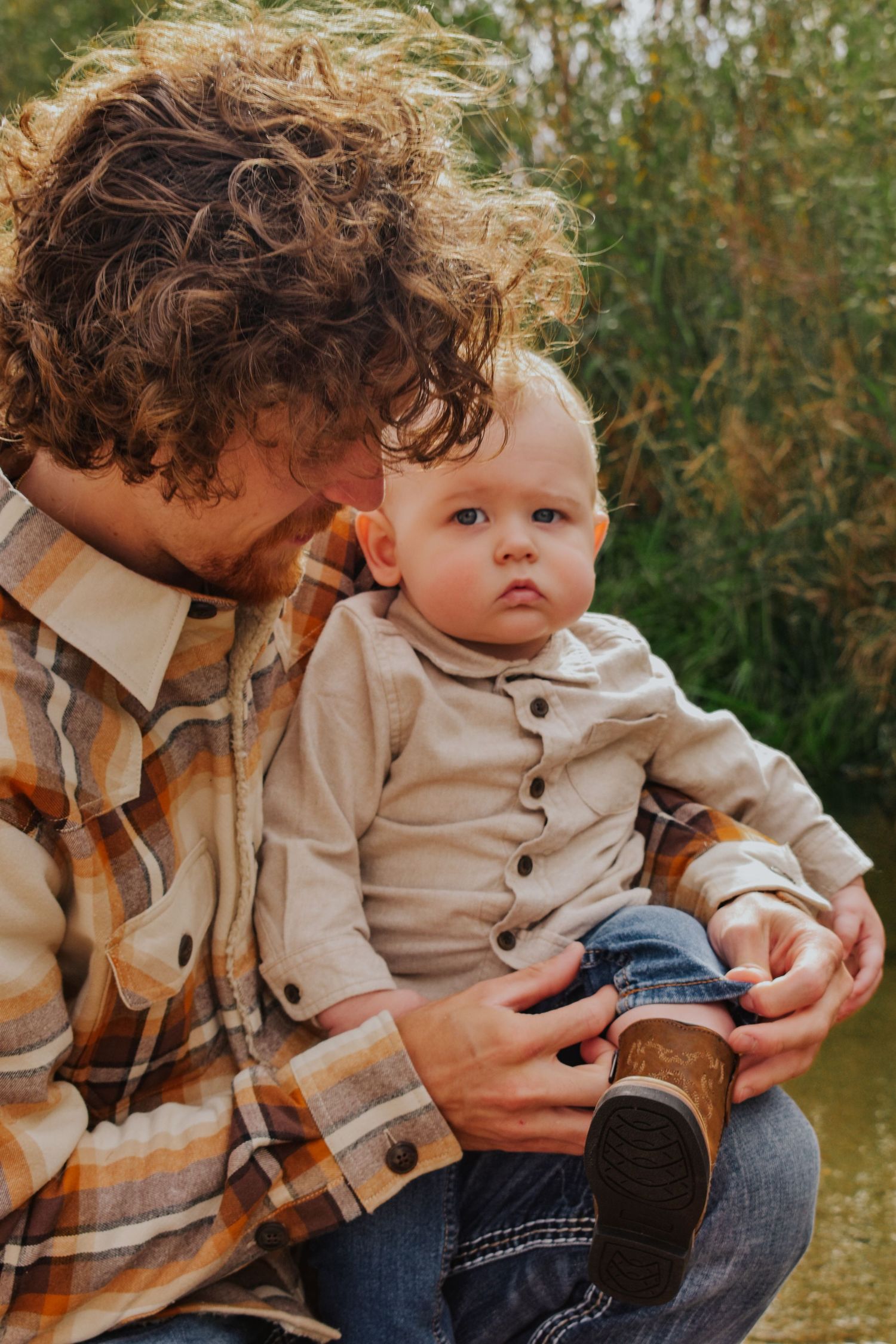 Curly-haired father in plaid shirt tenderly holding blonde baby boy outdoors near water.