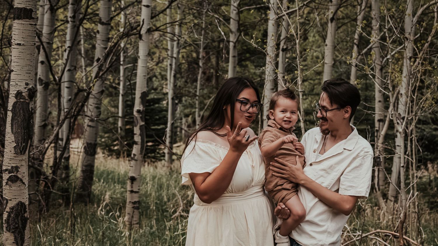 Two women smiling and holding a baby boy in a birch tree forest during a family photo session.