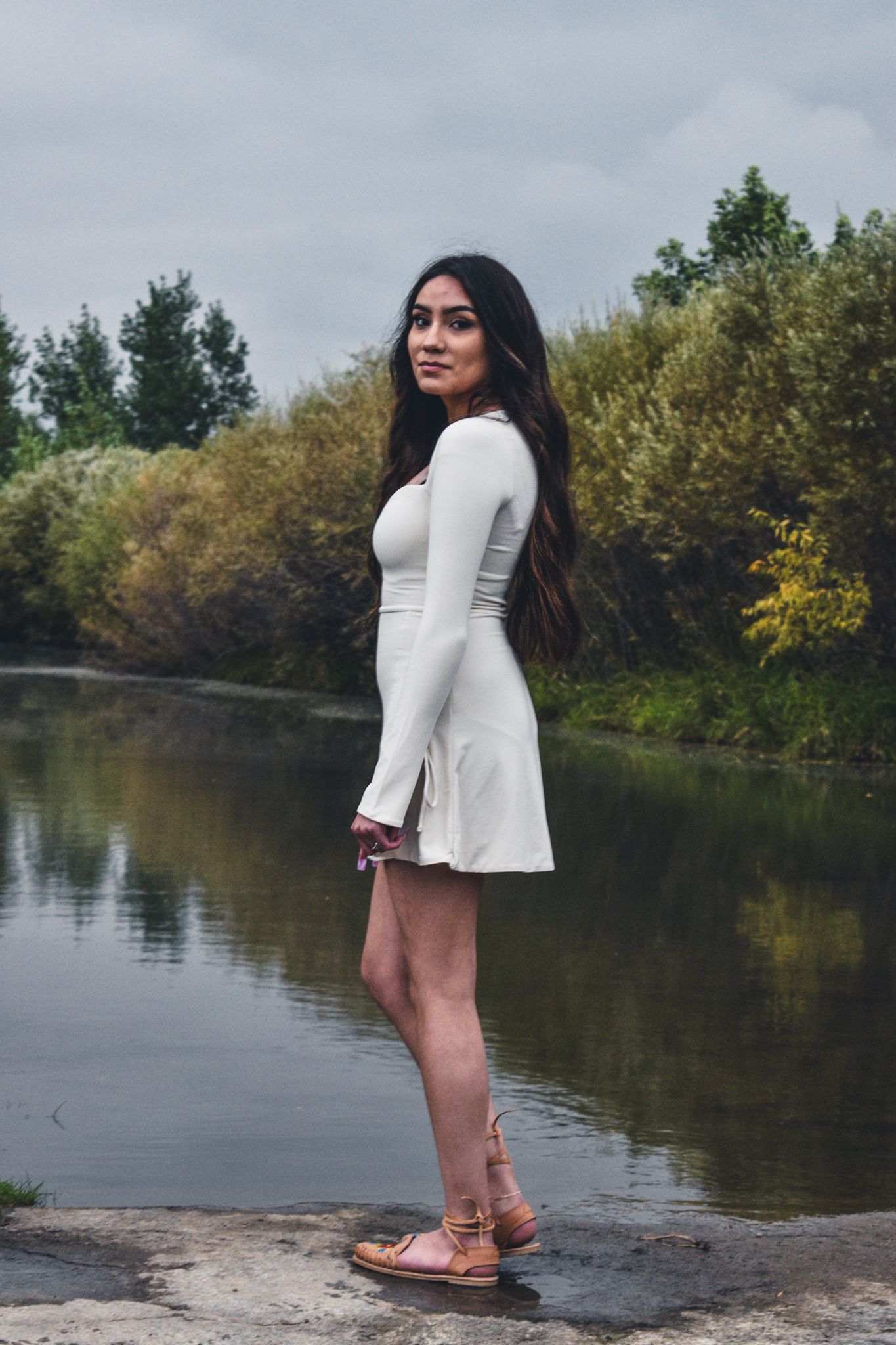 Young woman in white mini dress and sandals poses by a serene river surrounded by lush green trees for senior photos.