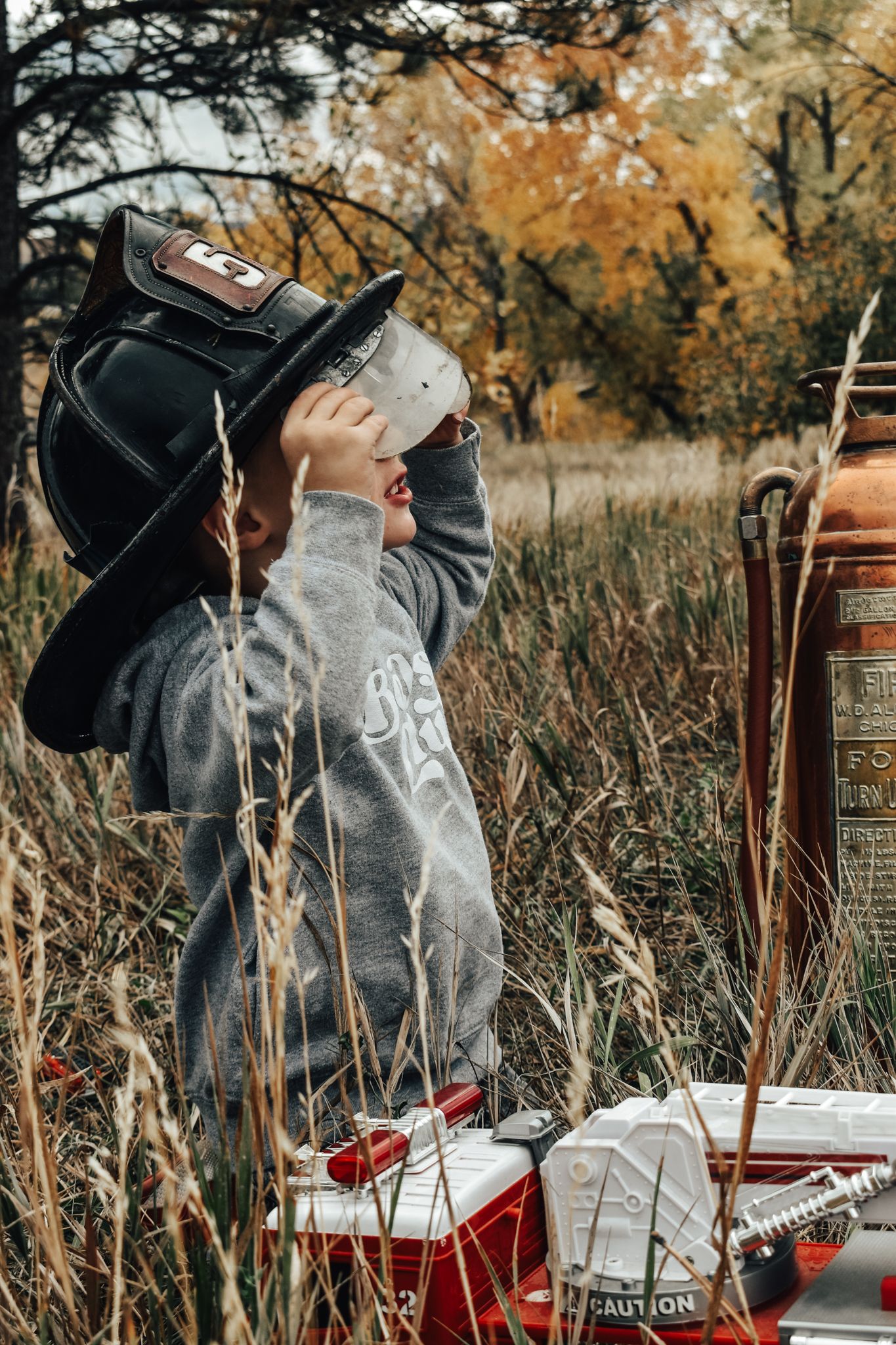 Young boy wearing a firefighter helmet explores tall grass in an autumn field near a fire truck.