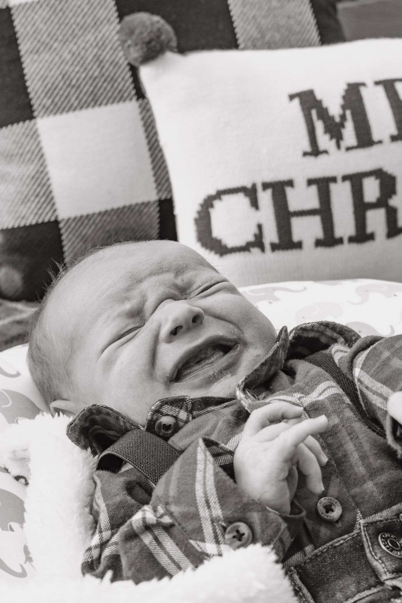 Crying newborn baby in plaid shirt with Christmas pillow in black and white photo. Family Photographer in Pflugerville, Tx