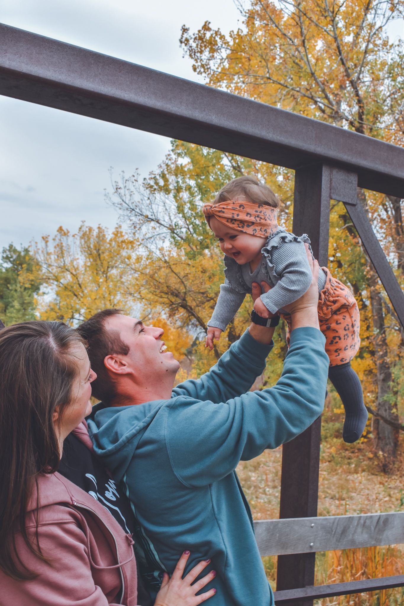 Parents lifting smiling baby girl on autumn bridge with colorful fall foliage background. Family Photographer in Pflugerville, Tx