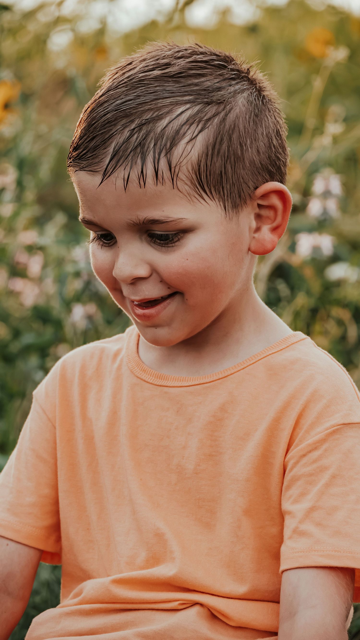 A young boy in an orange t-shirt smiles looking down, with colorful flowers in the background.