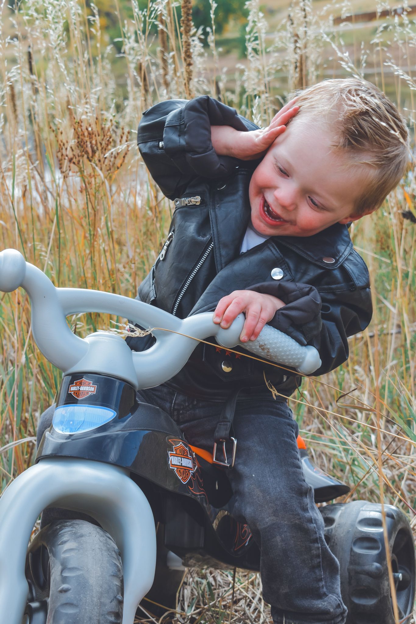 Toddler boy in Harley Davidson leather jacket sitting on toy motorcycle outdoors. Family Photographer in Pflugerville, Tx