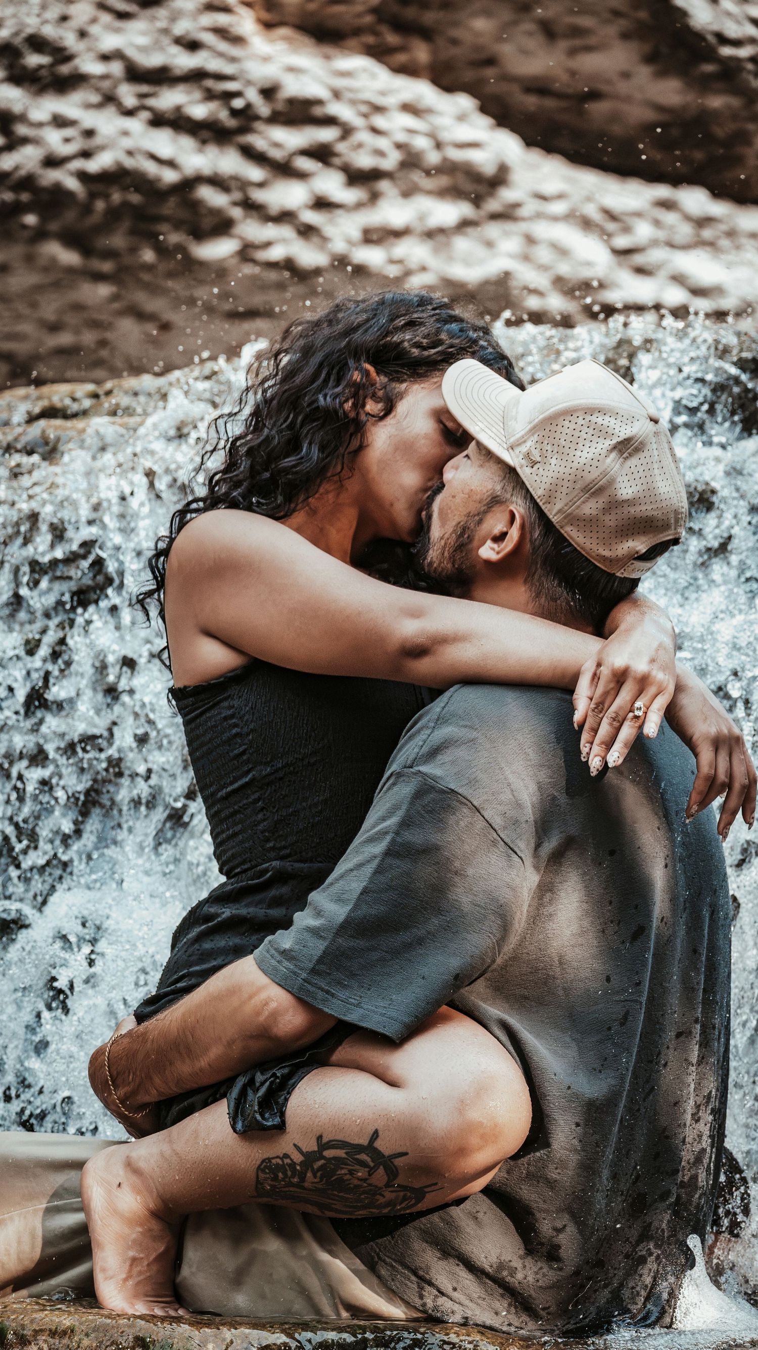 Couple kissing passionately by a waterfall, woman with curly hair embracing tattooed man in cap on rocks.