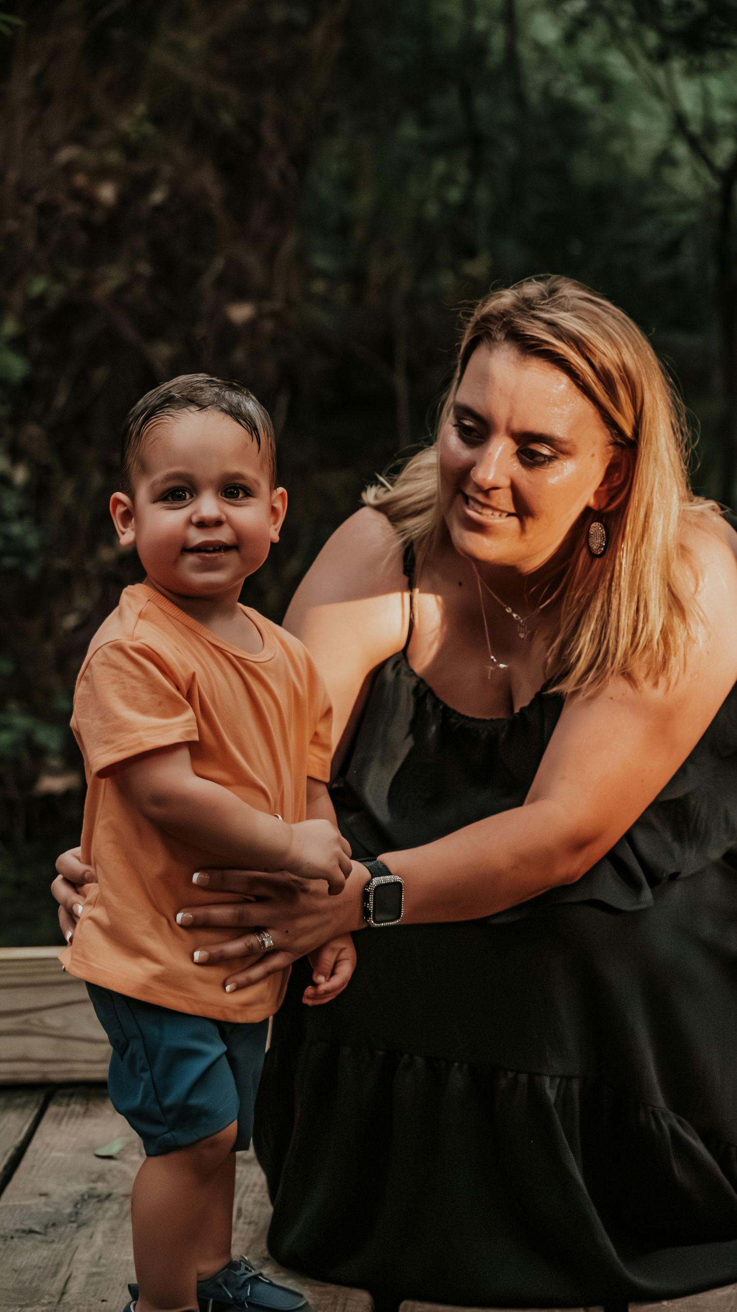 Smiling blonde mother in black dress crouches beside her young son in an orange shirt outdoors.
