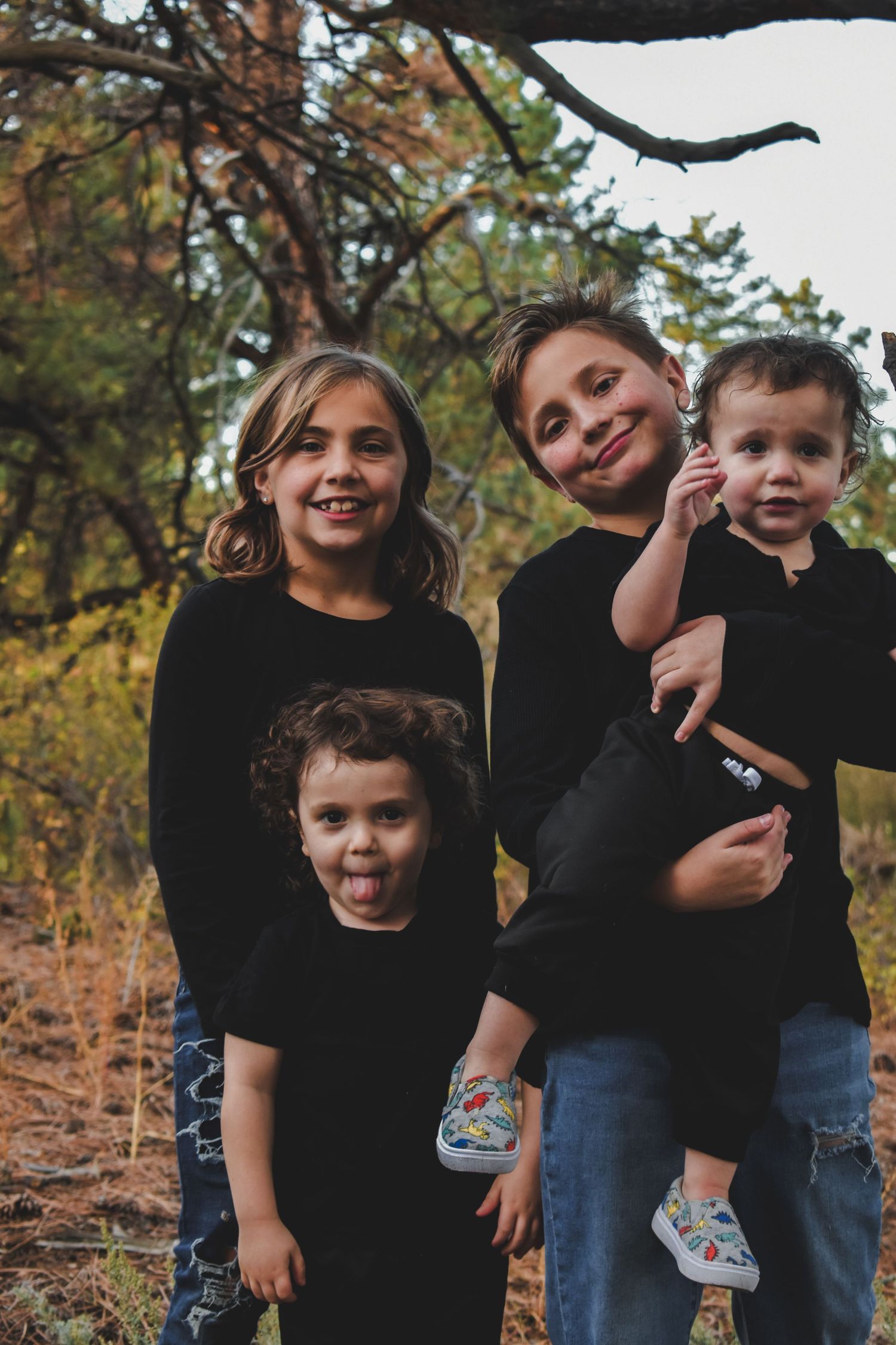Four siblings in black outfits posing playfully together in an autumn forest setting.