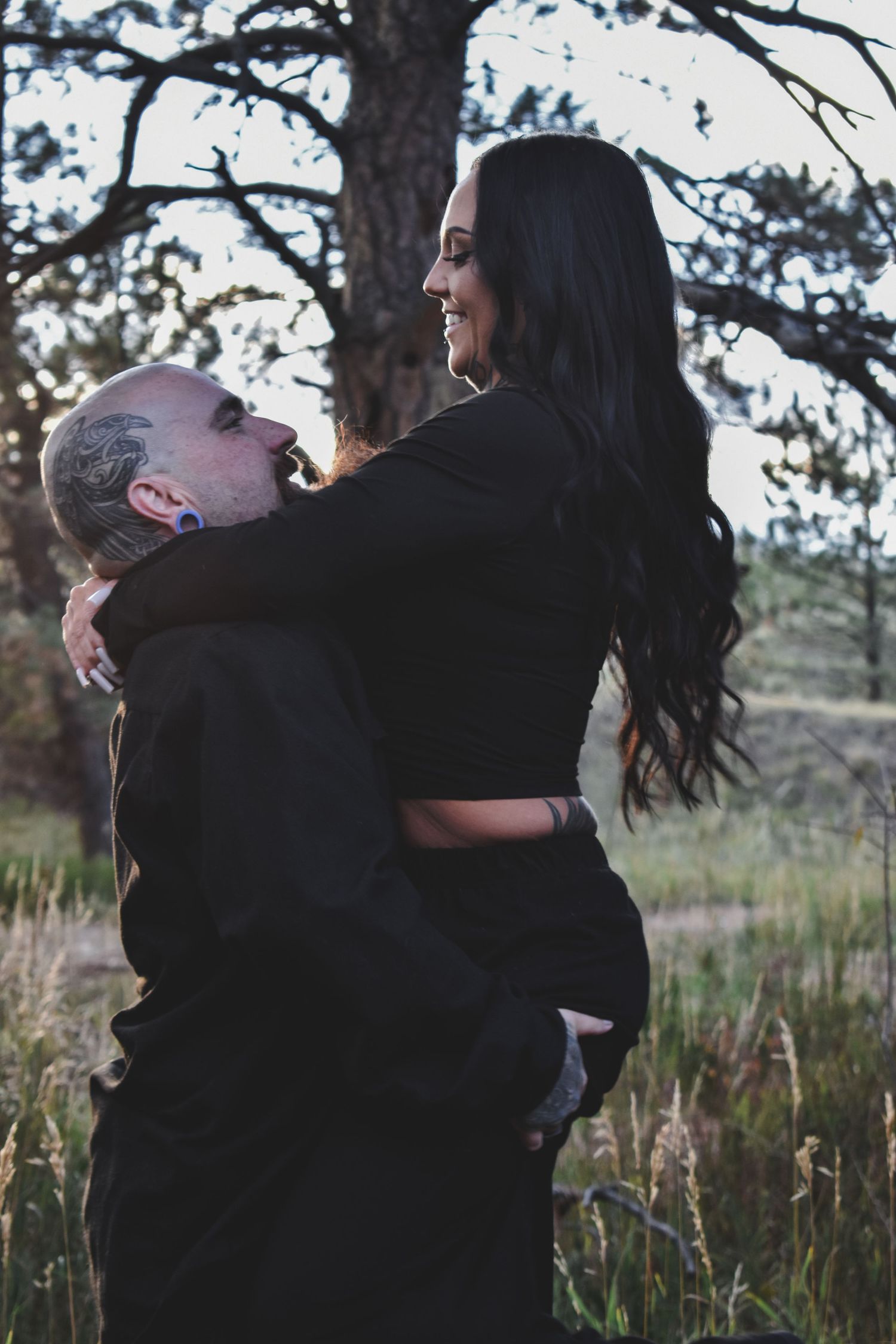 Man lifting laughing dark-haired woman in a romantic outdoor embrace during sunset in a grassy field.