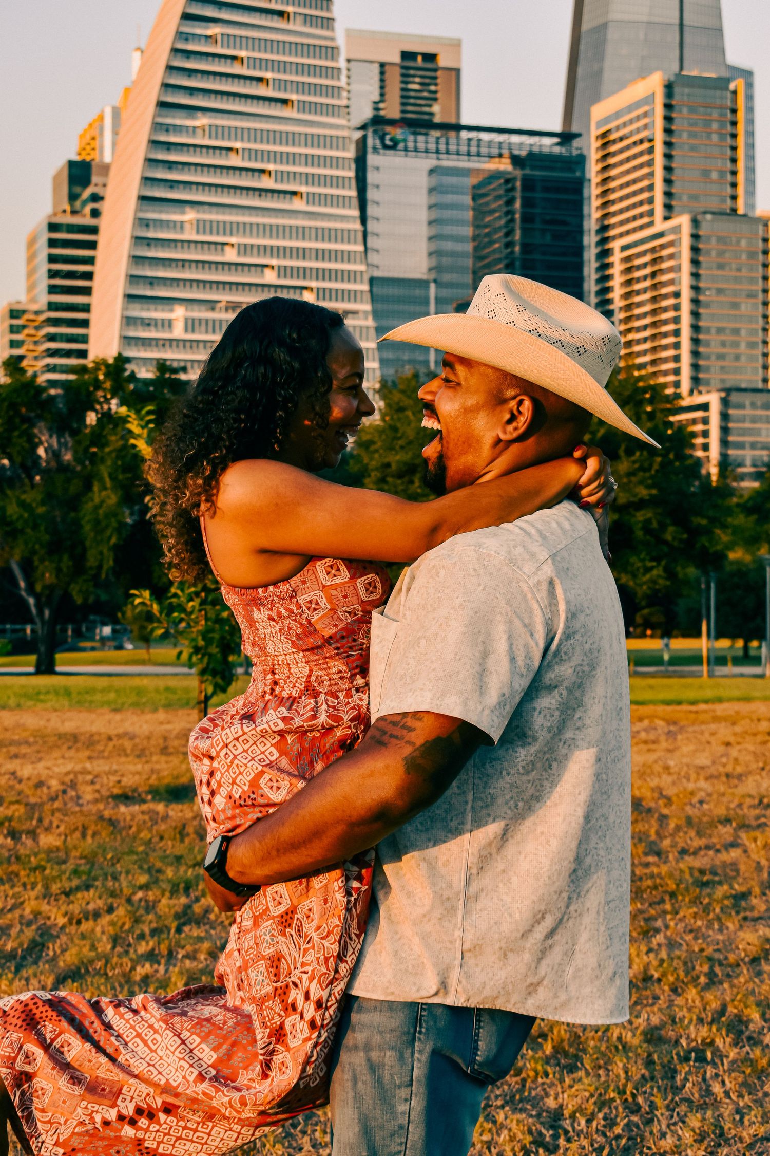 Man in cowboy hat lifting woman in floral dress while embracing in a park at golden hour.