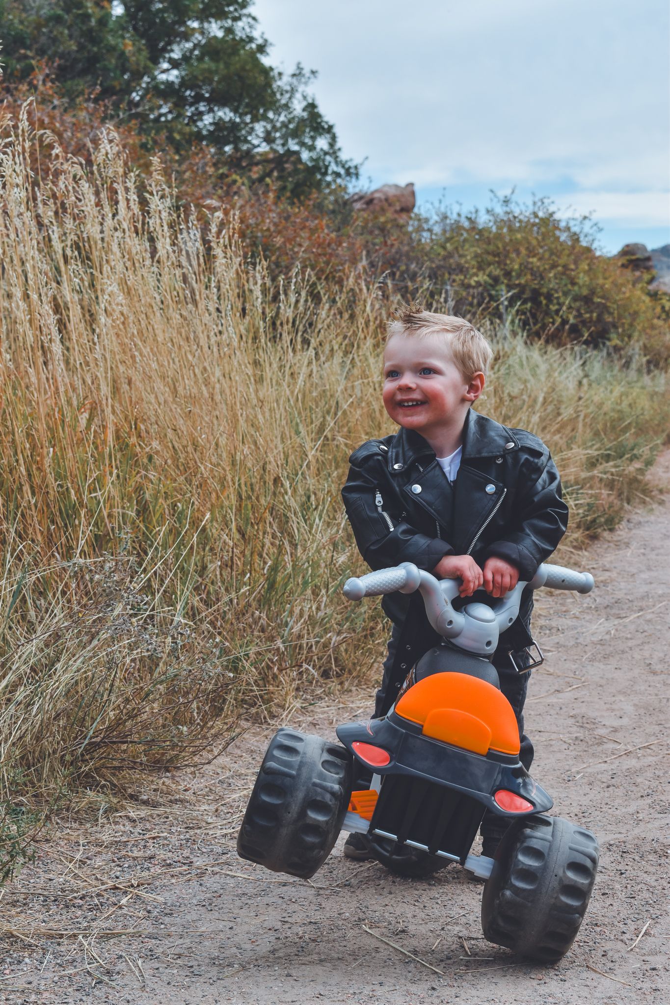 Laughing toddler in black leather jacket riding orange and black toy tricycle on dirt trail.