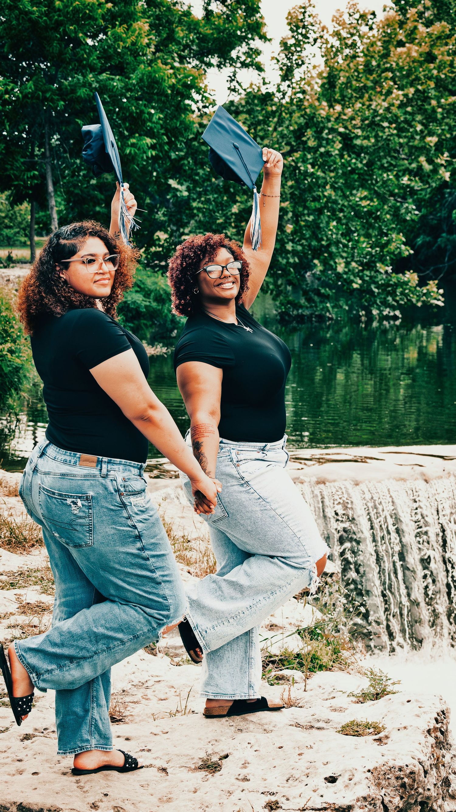Two smiling female graduates toss caps near a scenic waterfall wearing black tops and jeans for senior photos.