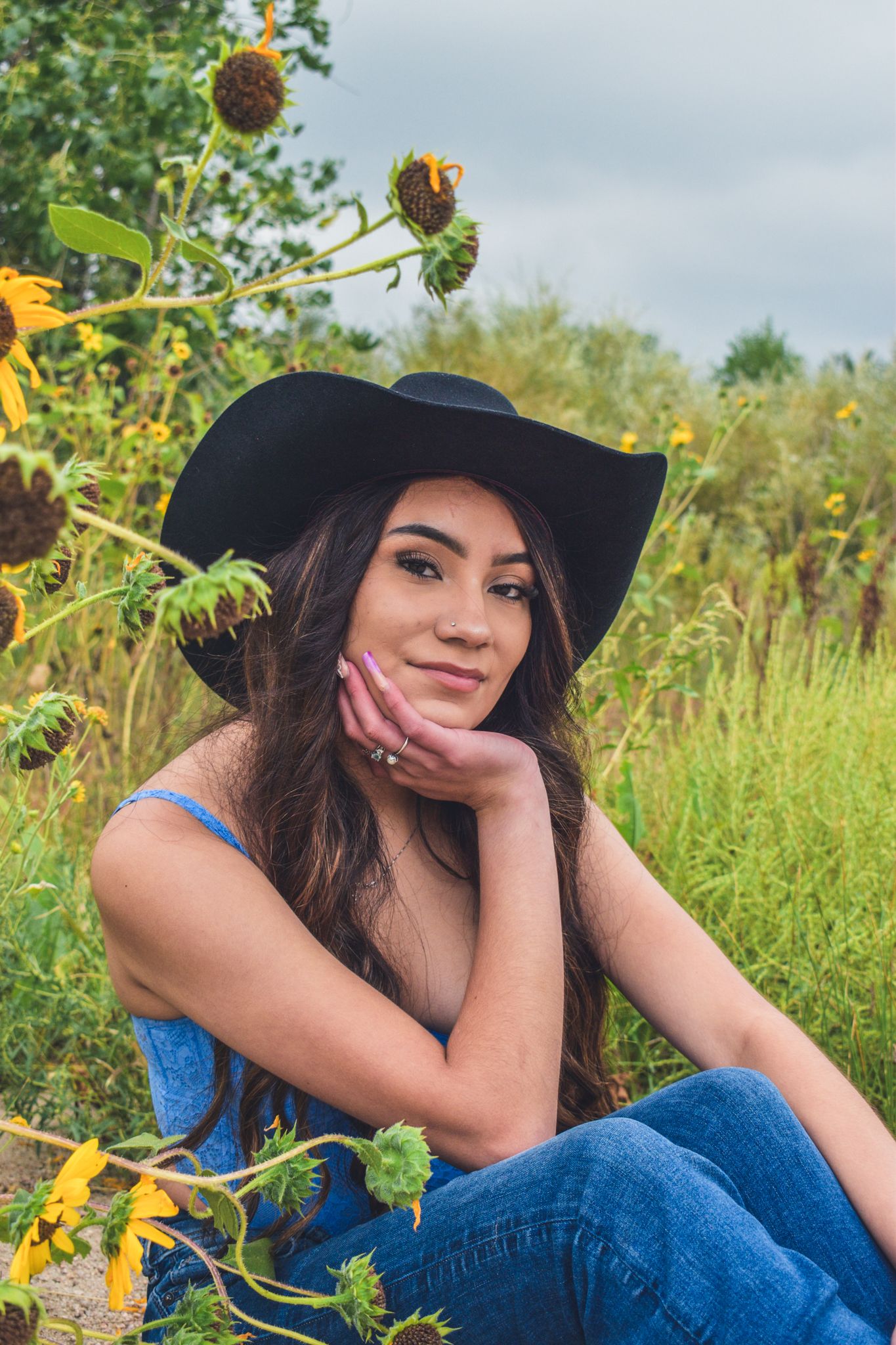 Young woman in black wide-brim hat sits among sunflowers in a field, resting chin on hand for senior photos.