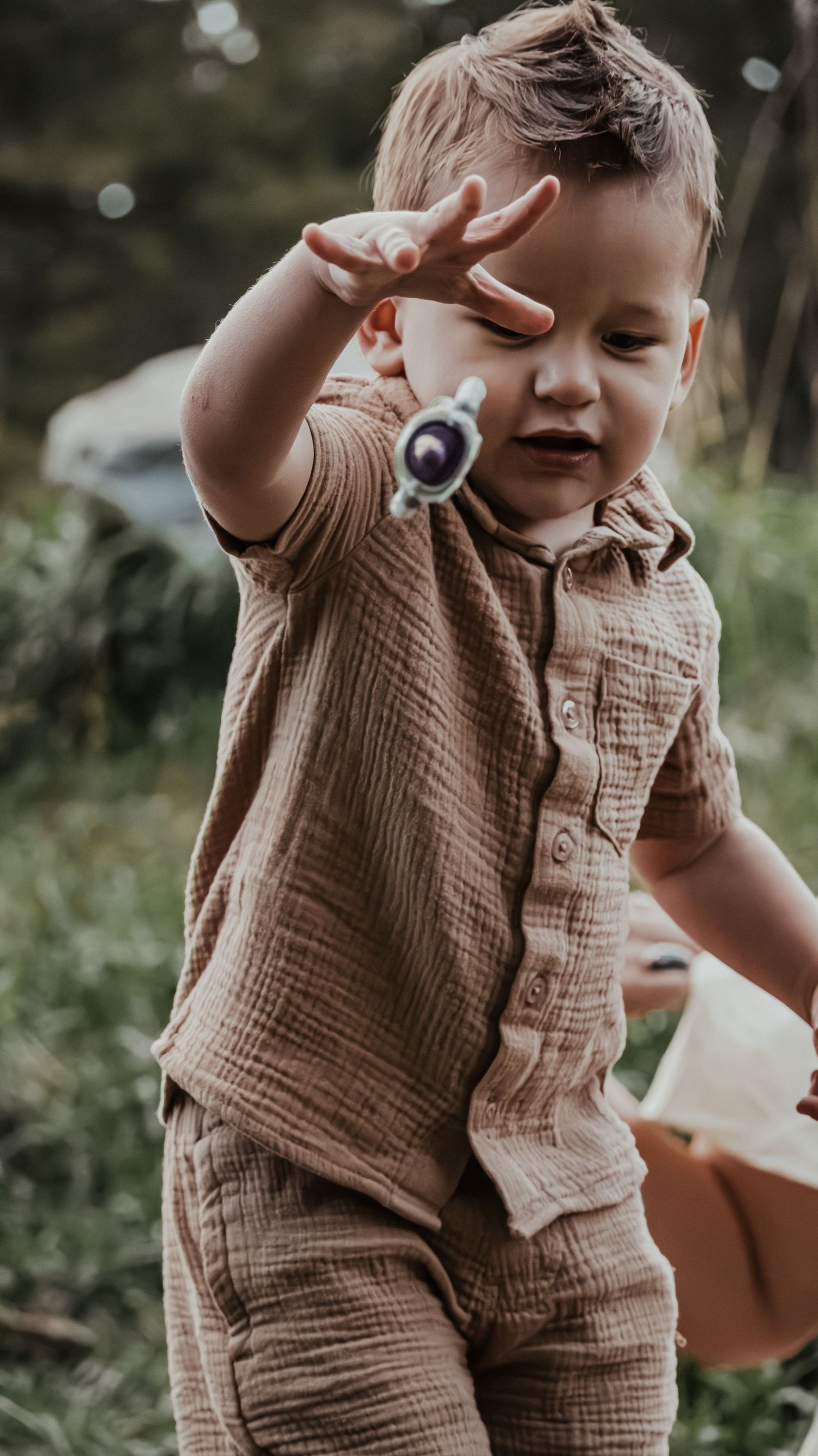 Curious toddler boy in brown linen outfit playing with small toy outdoors in grassy park setting.