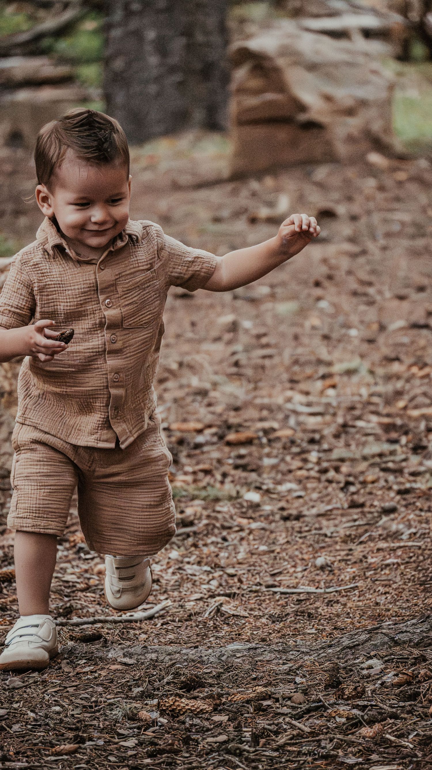 Happy toddler boy in tan linen outfit runs joyfully along a woodland forest path.