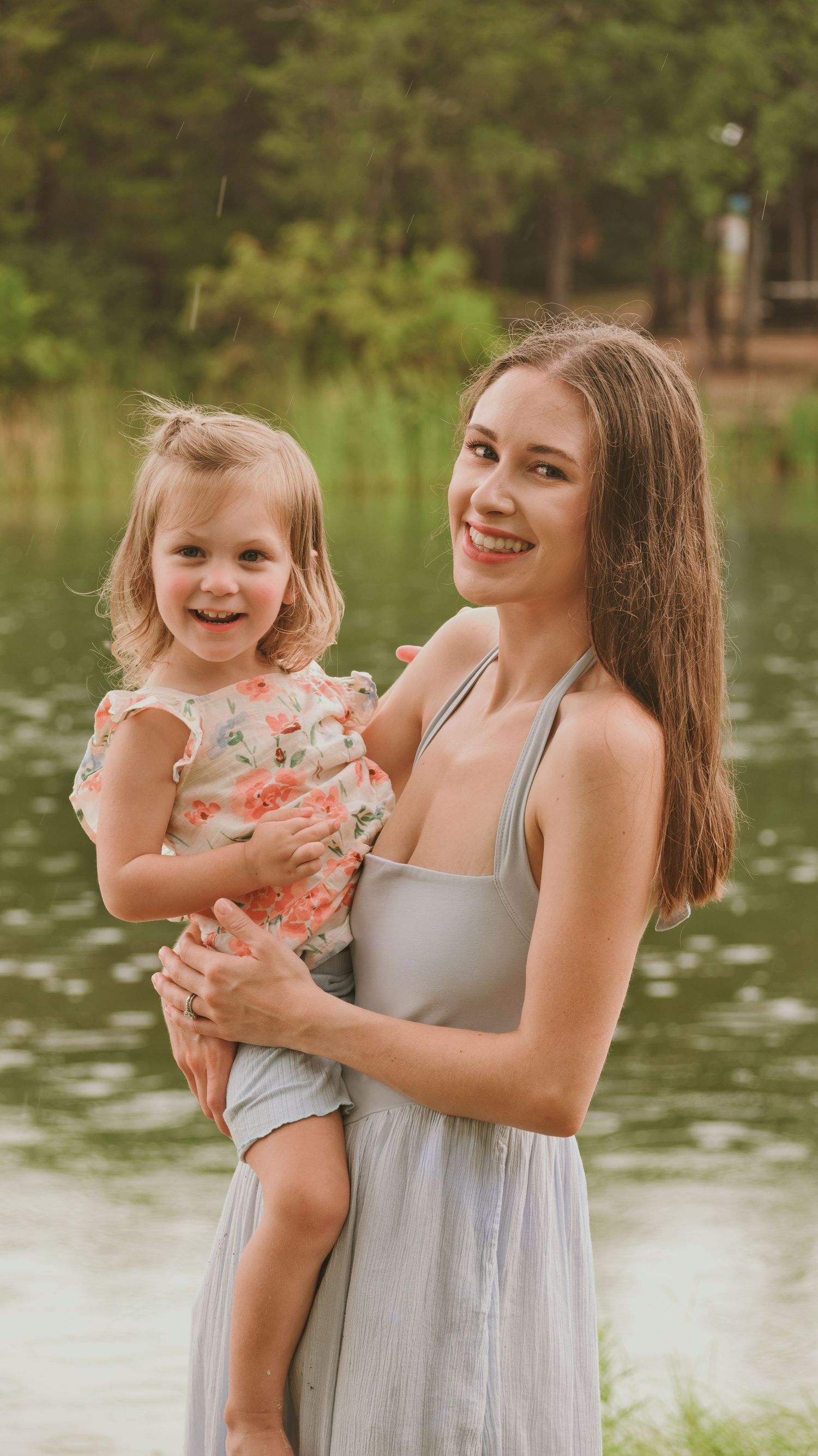 Smiling mother in gray dress holds her happy blonde toddler daughter near a peaceful lake.