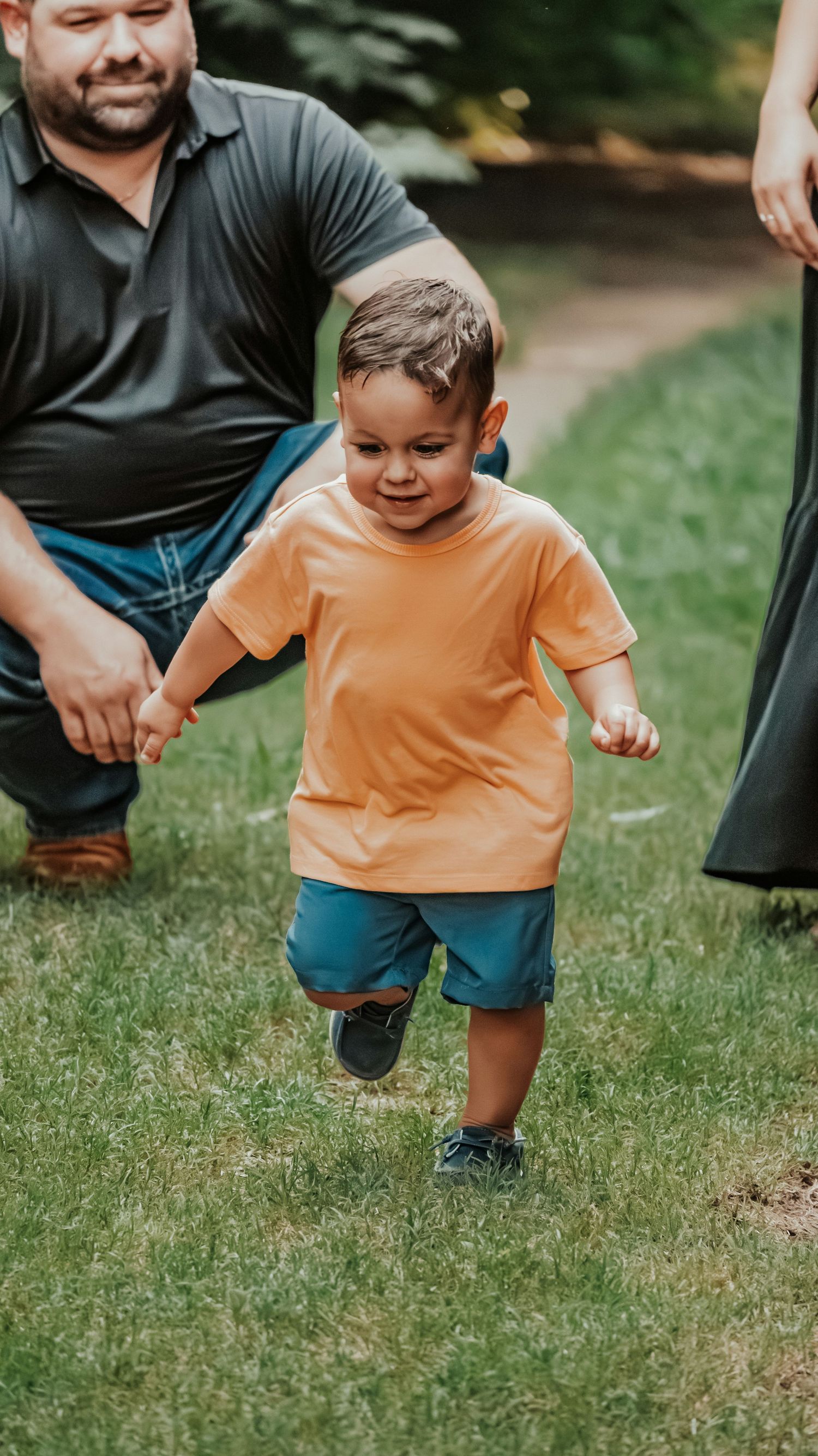 Toddler boy in orange shirt and blue shorts running on grass while father kneels behind him outdoors.