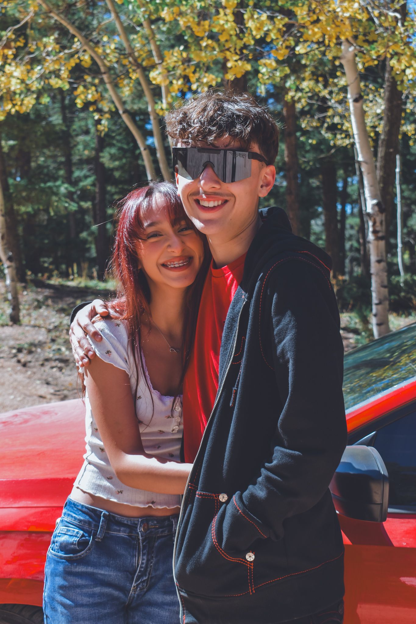 Smiling young couple embracing near a red car in a sunny autumn forest setting for senior photos.