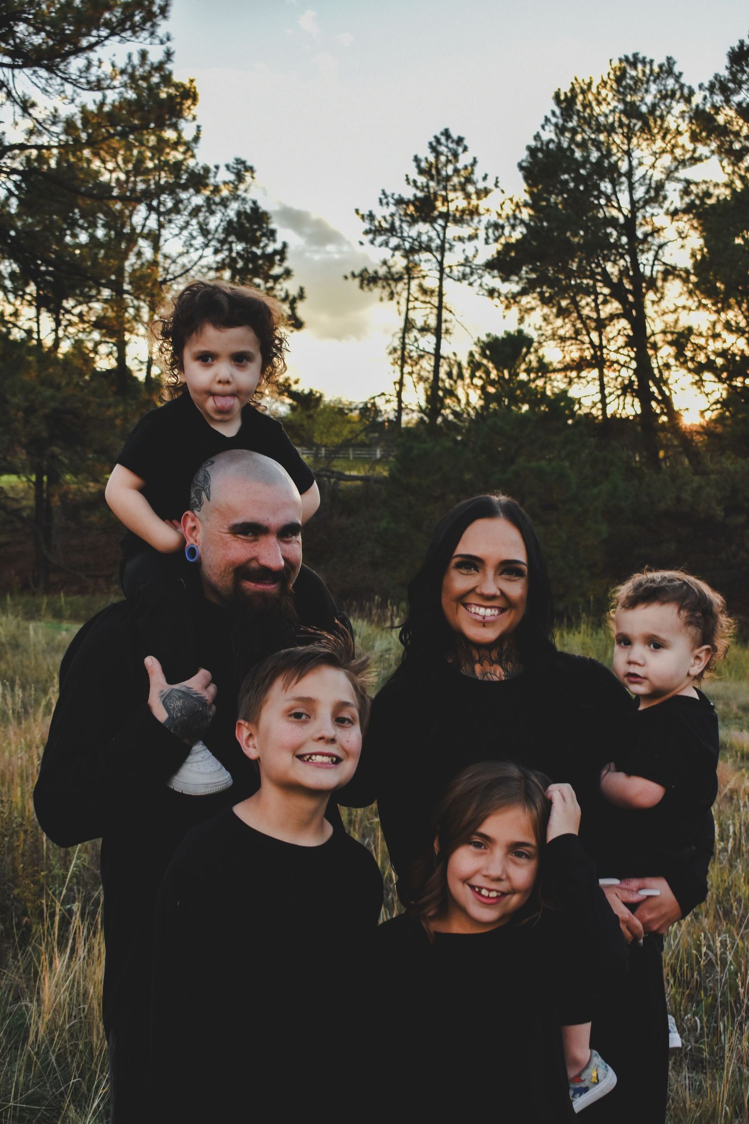 Family of six in matching black outfits posing outdoors at sunset with pine trees in the background. Family Photographer in Pflugerville, Tx