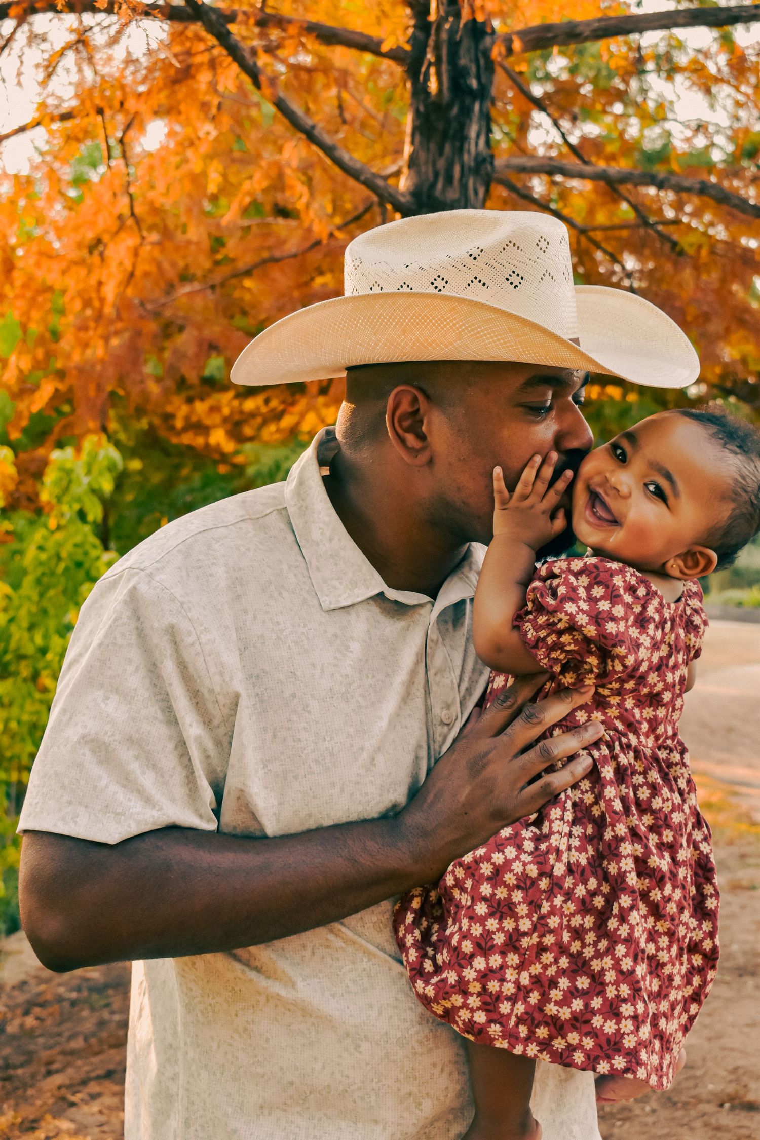 A loving father in a cowboy hat kisses his baby daughters cheek in a vibrant autumn park.