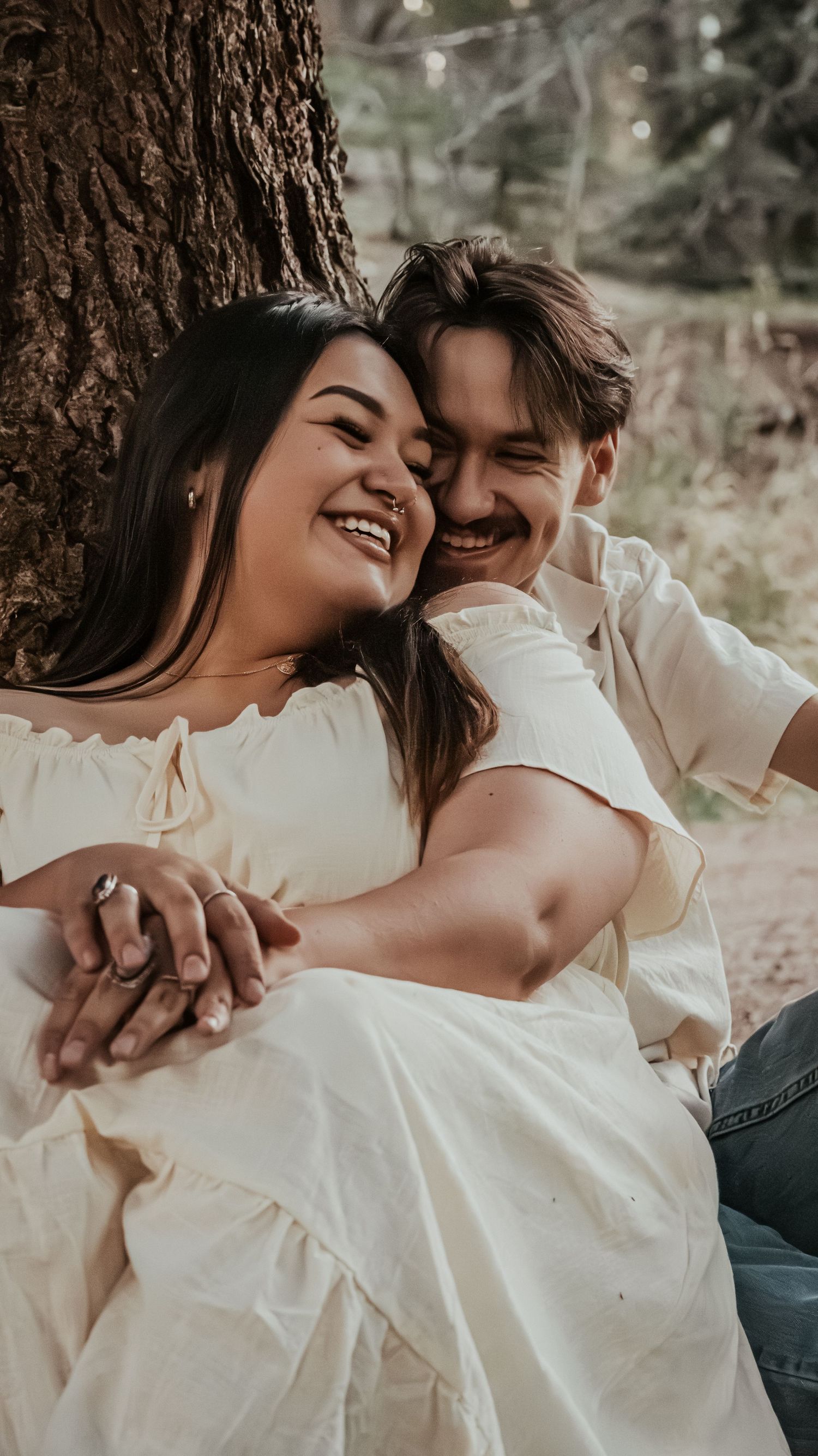 A smiling couple embraces warmly while sitting together against a tree trunk outdoors.