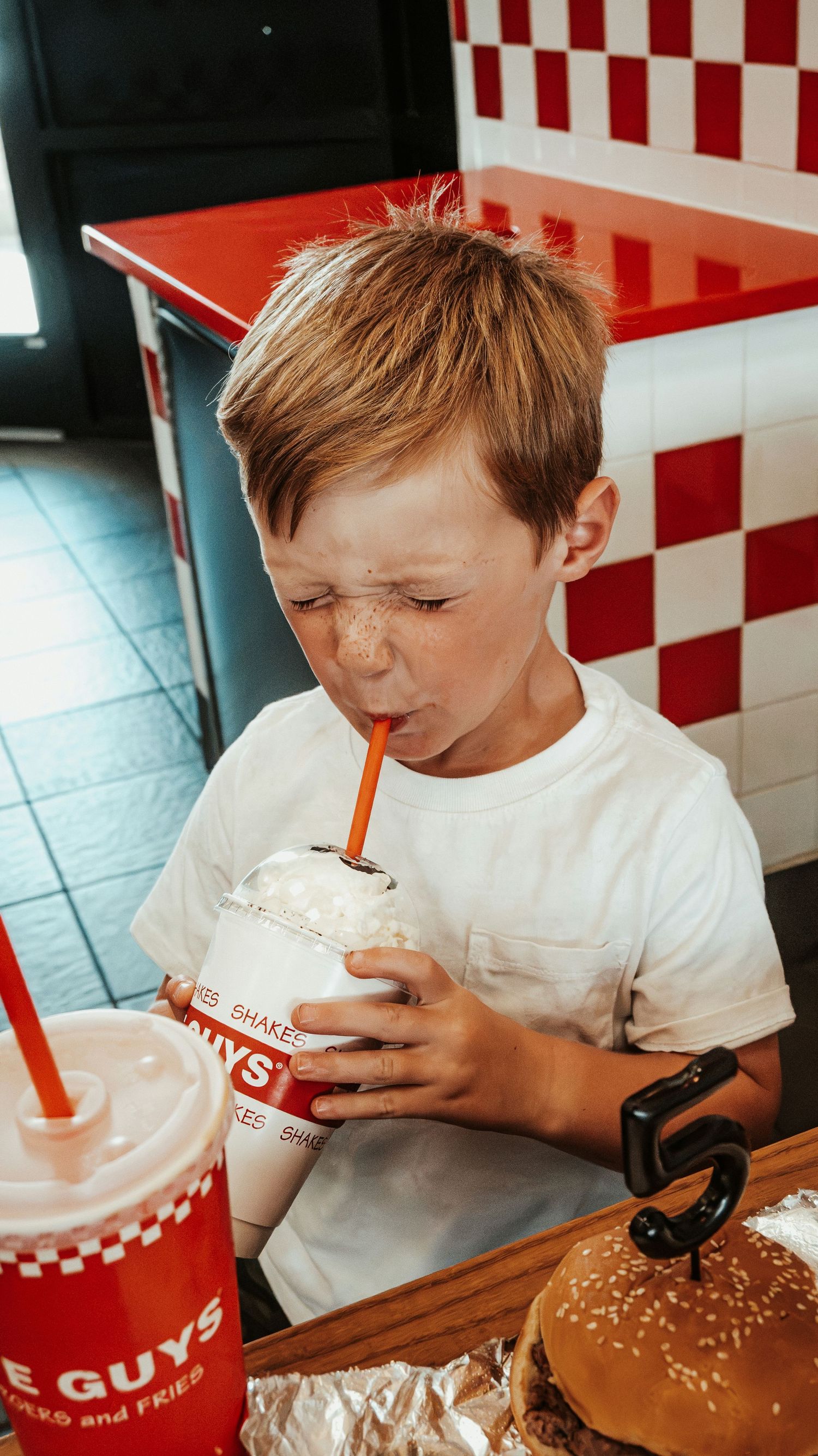Boy sipping a milkshake at Five Guys burger restaurant with red checkered decor.