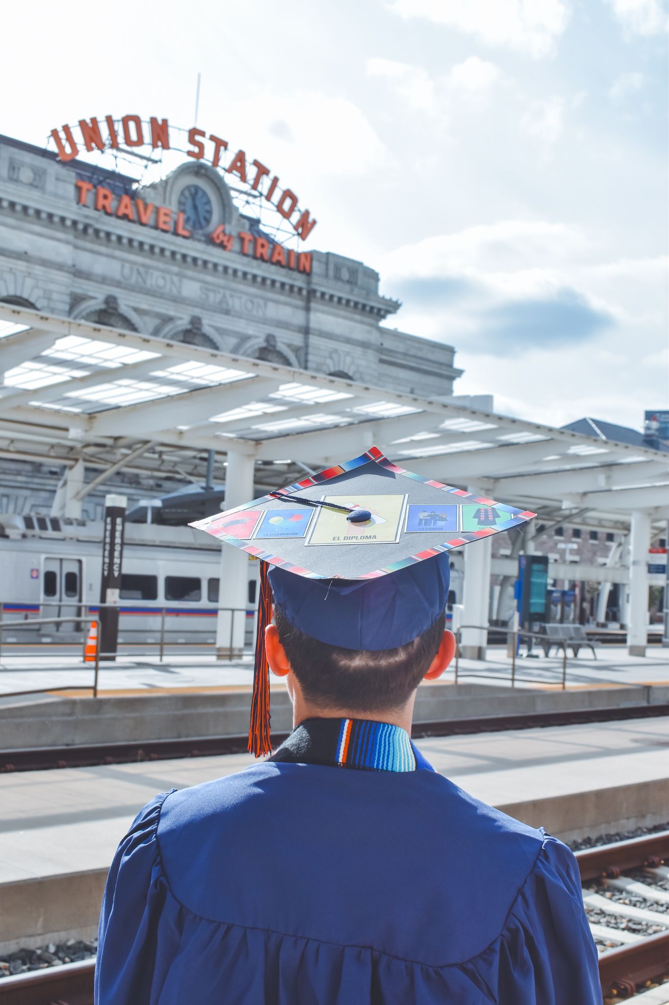 Graduate in decorated mortarboard cap viewed from behind at Denver Union Station train platform for senior photos.