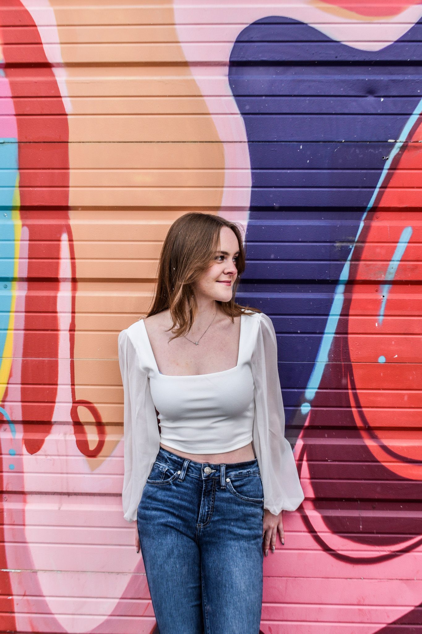 Young woman in white sheer-sleeve crop top and jeans posing in front of colorful urban graffiti mural for senior photos.
